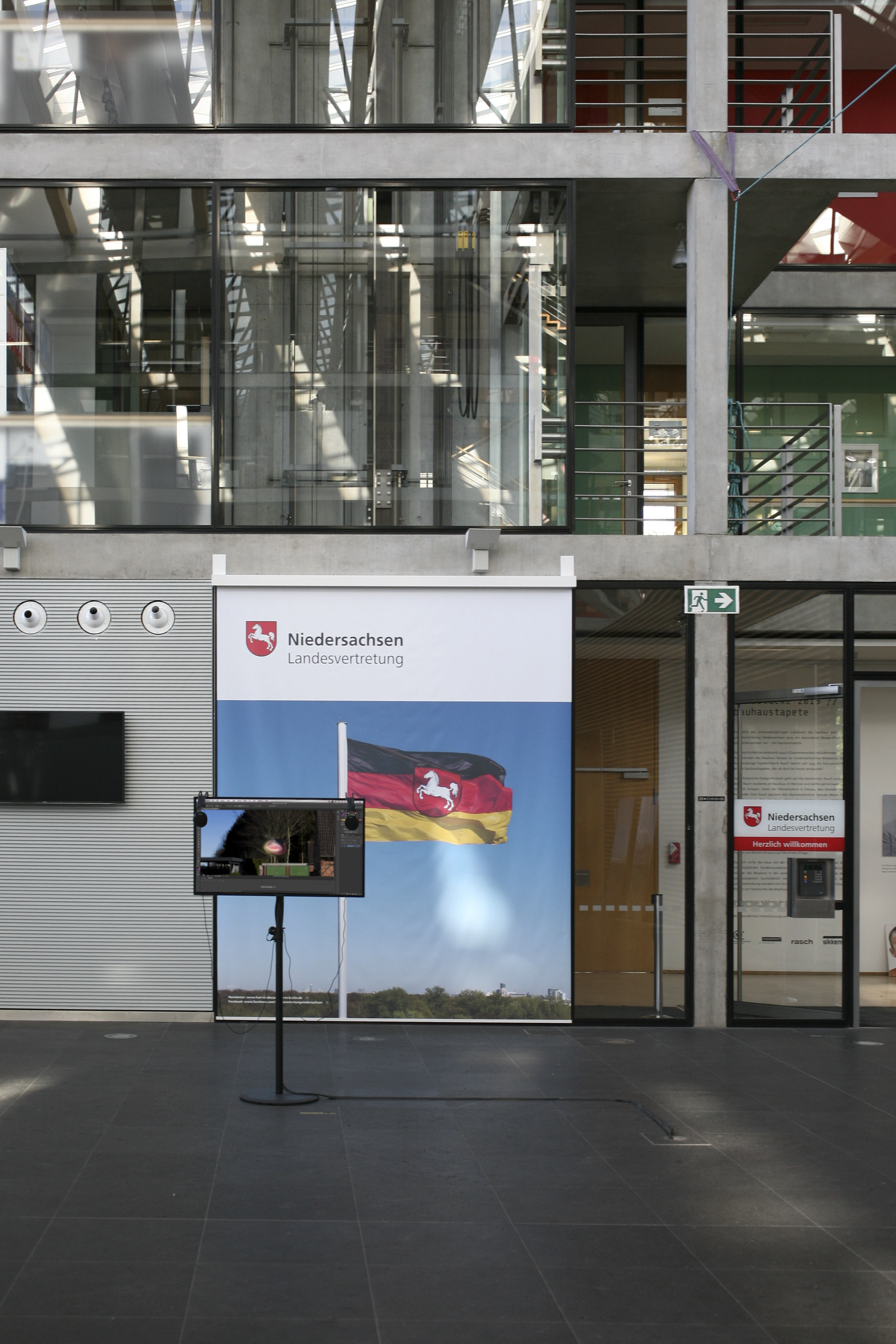 Installation view in a glass atrium: a monitor on a stand in front of a banner showing the flag of Lower Saxony and the words 'Niedersachsen Landesvertretung'. Angels Tutorial (2018) by Nickii Schamborski.