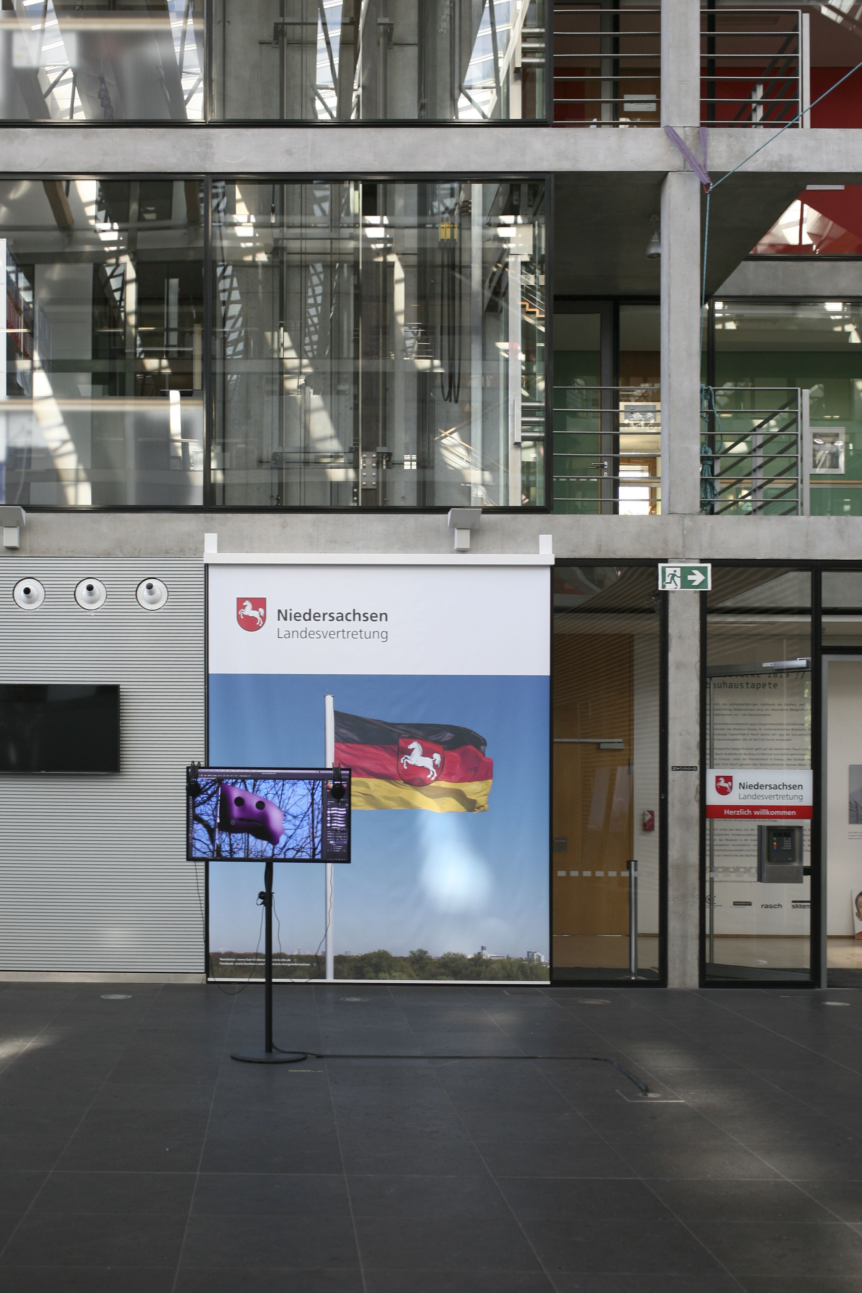 Installation view in the glass atrium: the monitor in front of the Lower Saxony banner, showing a different frame of the video. Angels Tutorial (2018) by Nickii Schamborski.