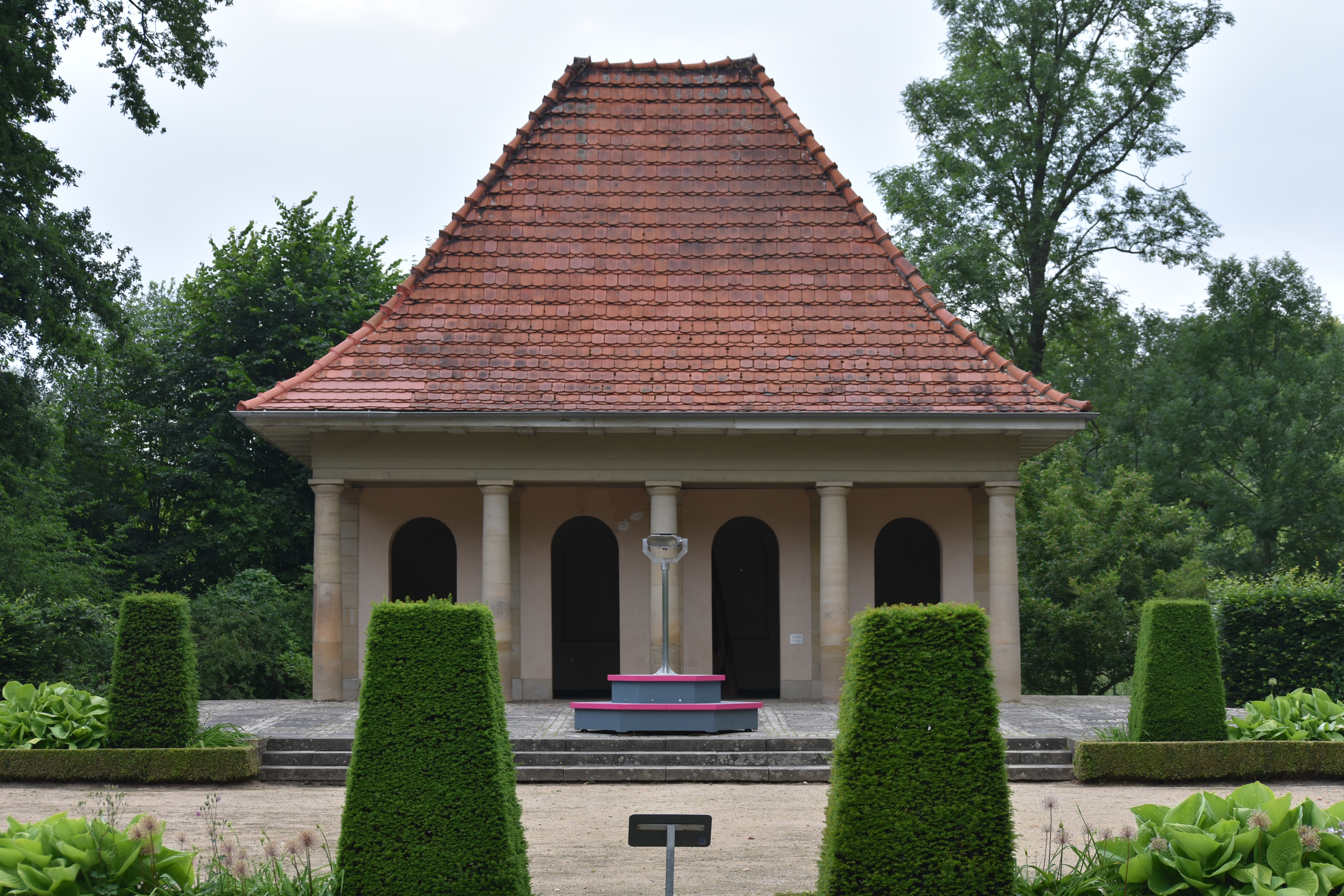A garden pavilion with a red tiled roof and columns, framed by trimmed yew hedges; the viewing device stands on a hexagonal plinth in front of it. Instrument of Public Critique (2018) by Nickii Schamborski.