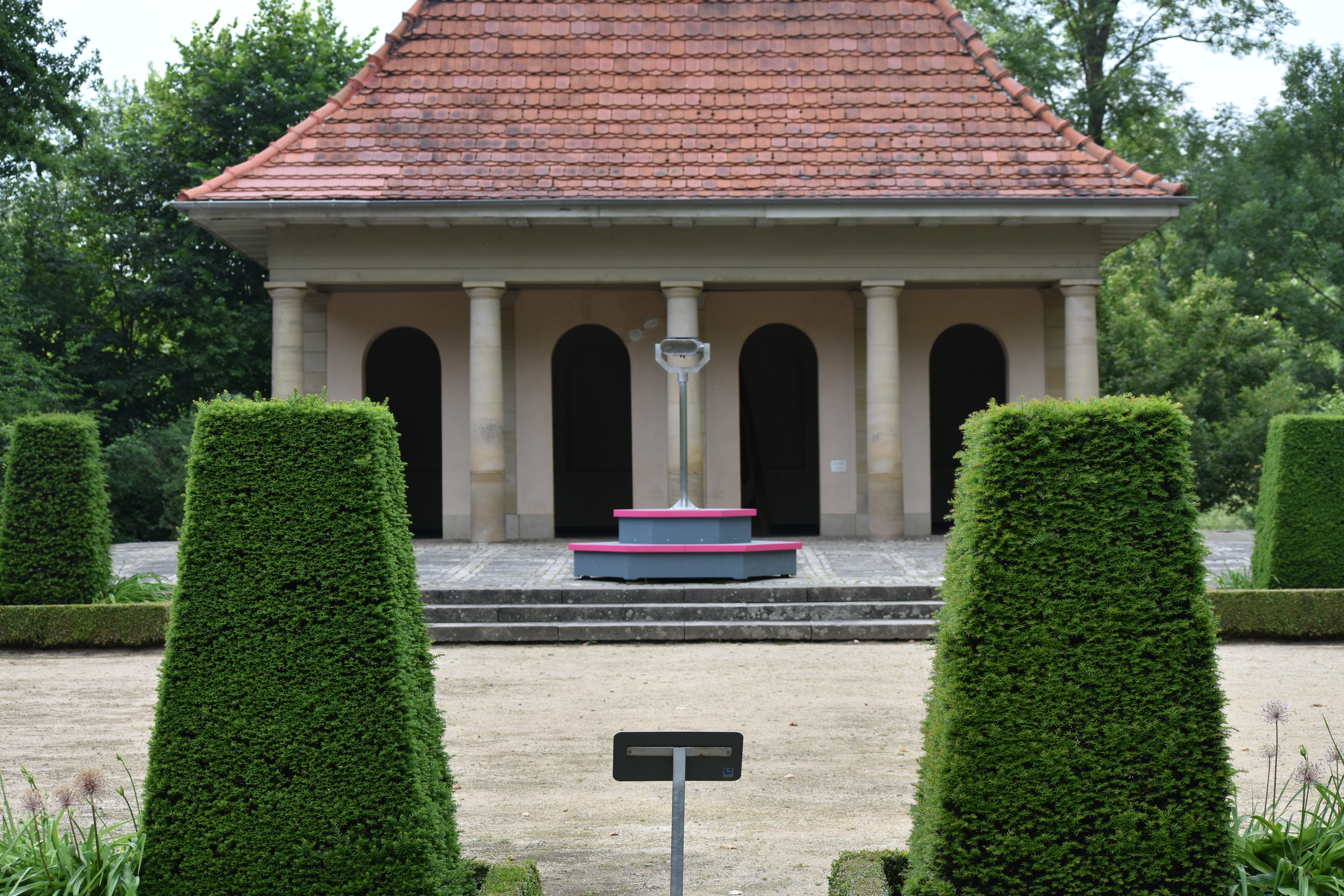 Closer view of the pavilion: the binocular viewing device on its grey and magenta plinth between trimmed hedges. Instrument of Public Critique (2018) by Nickii Schamborski.