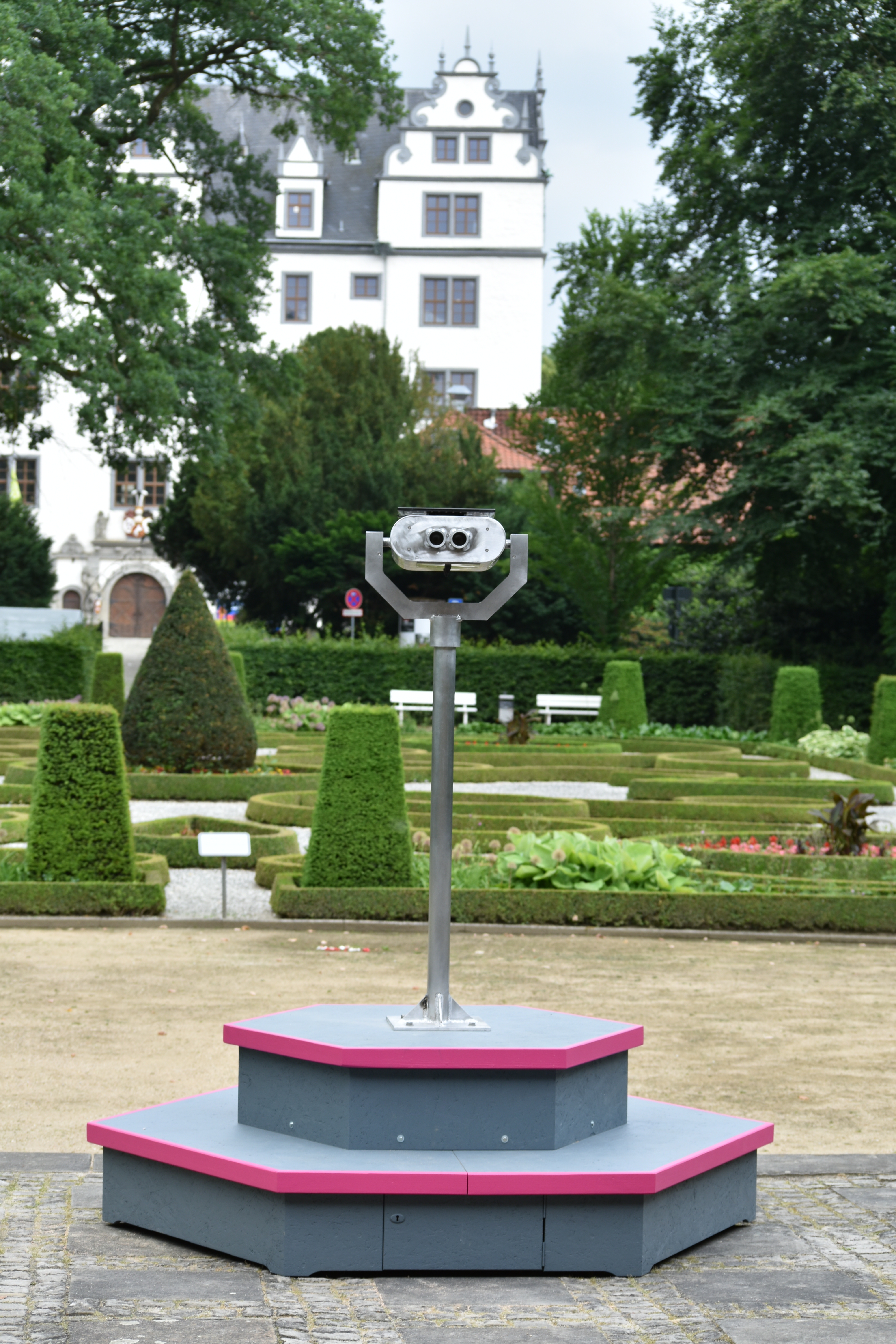 The sculpture: a metal binocular viewer on a pole, mounted on a stepped hexagonal plinth with magenta edges, in a formal garden with a white castle behind. Instrument of Public Critique (2018) by Nickii Schamborski.