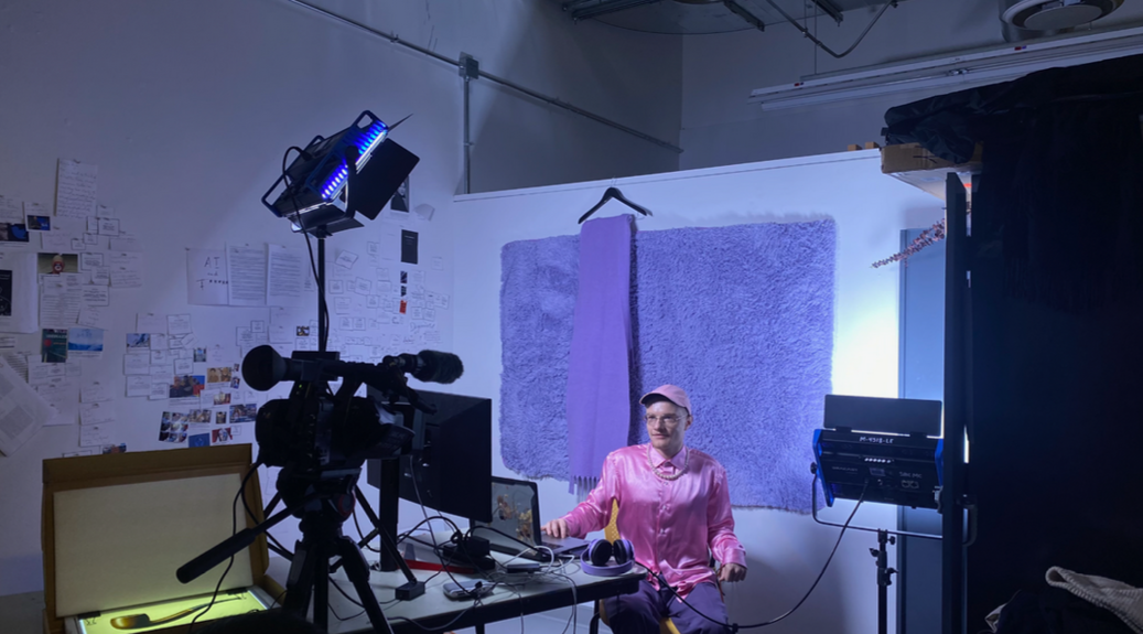 A person in a pink satin shirt and pink cap sits at a desk between studio lights and a camera, in front of a lilac fluffy backdrop. Performance: Nickii AI (2026) by Nickii Schamborski.