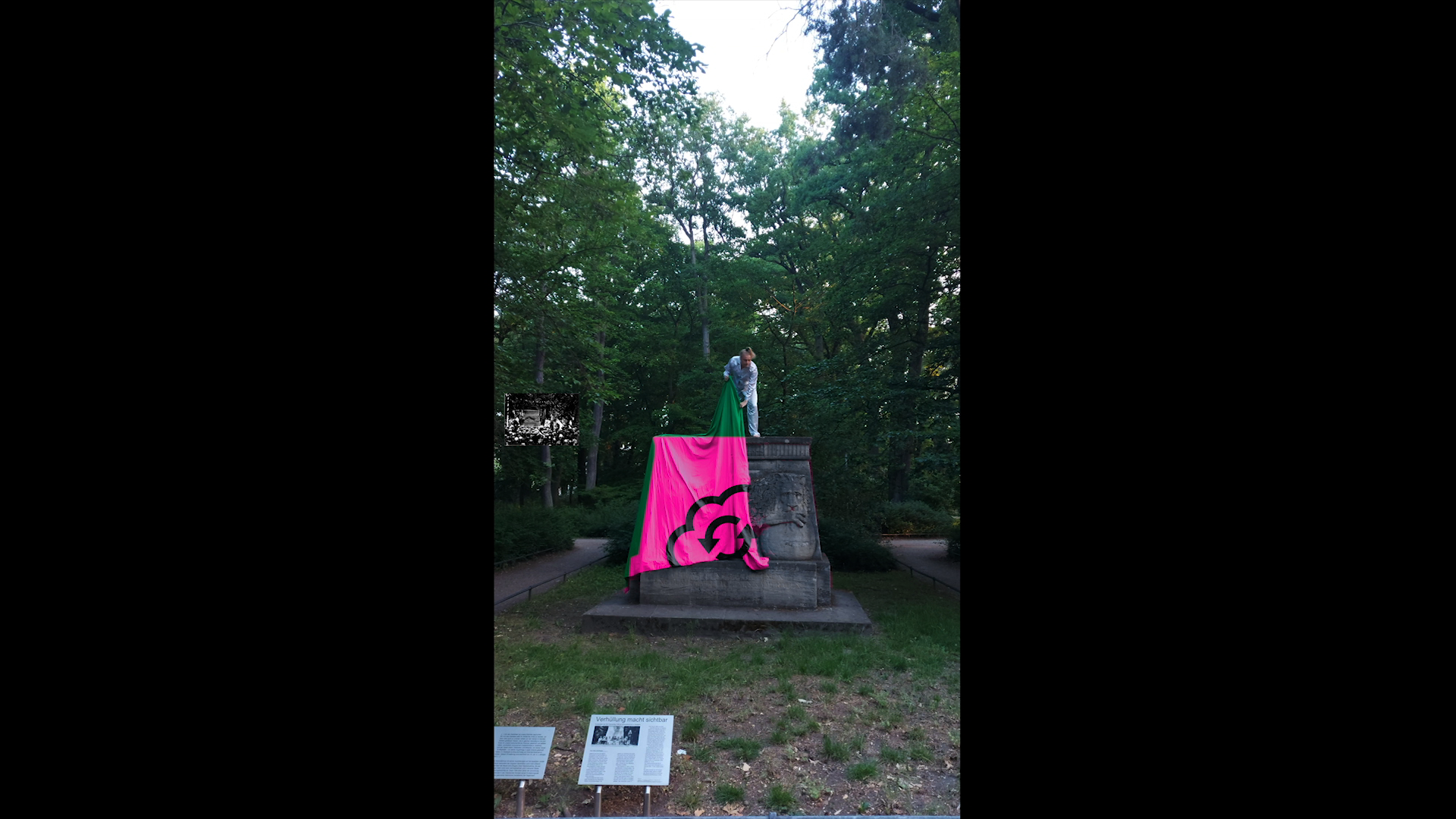 Phone view of the stone monument in the park, covered with a pink cloth carrying a black drawing; a person stands on top. Testing Trixi (2020) by Nickii Schamborski.