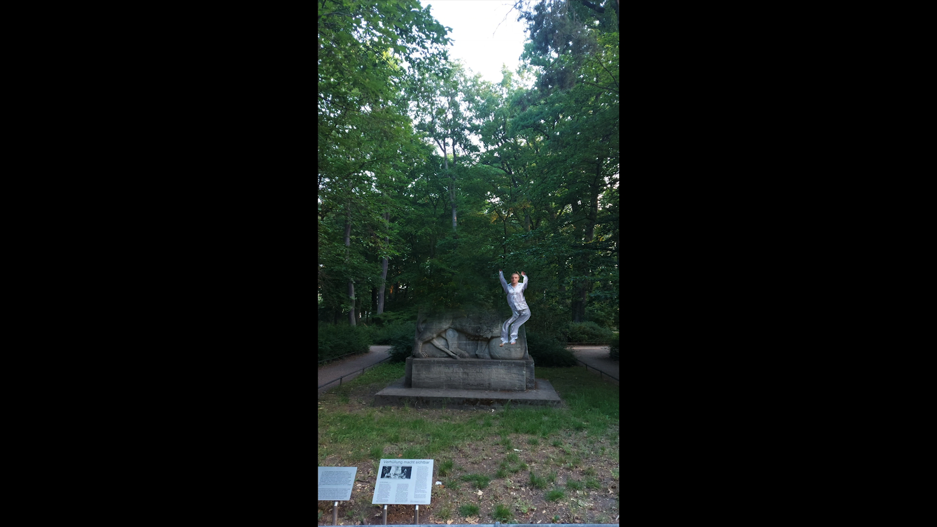 Phone view: a person stands with raised arms on top of the stone monument in the park. Testing Trixi (2020) by Nickii Schamborski.