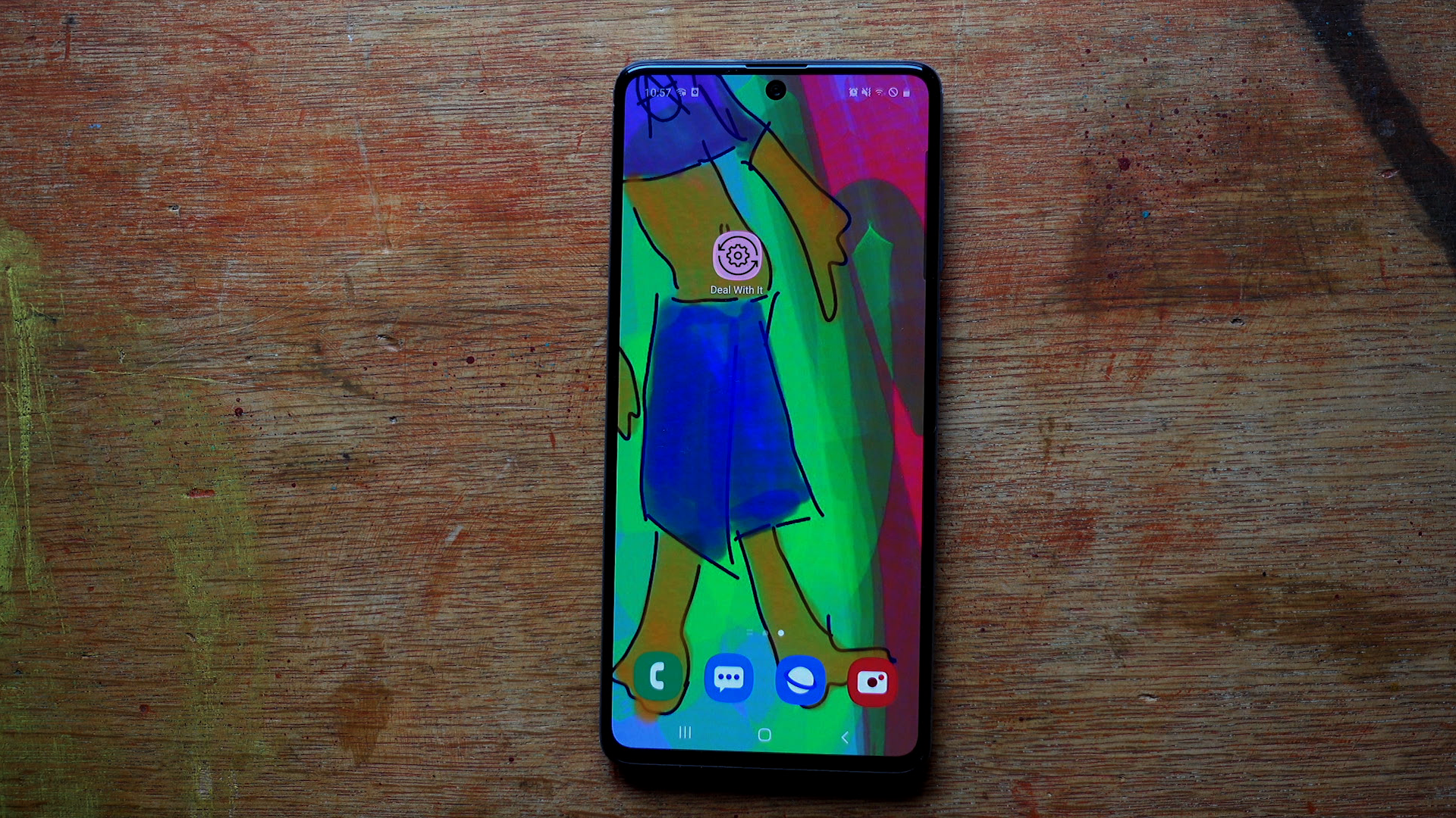 A smartphone lies on a paint-stained wooden surface; its screen shows a colourful drawn figure in a blue skirt behind the app icons. Testing Trixi (2020) by Nickii Schamborski.