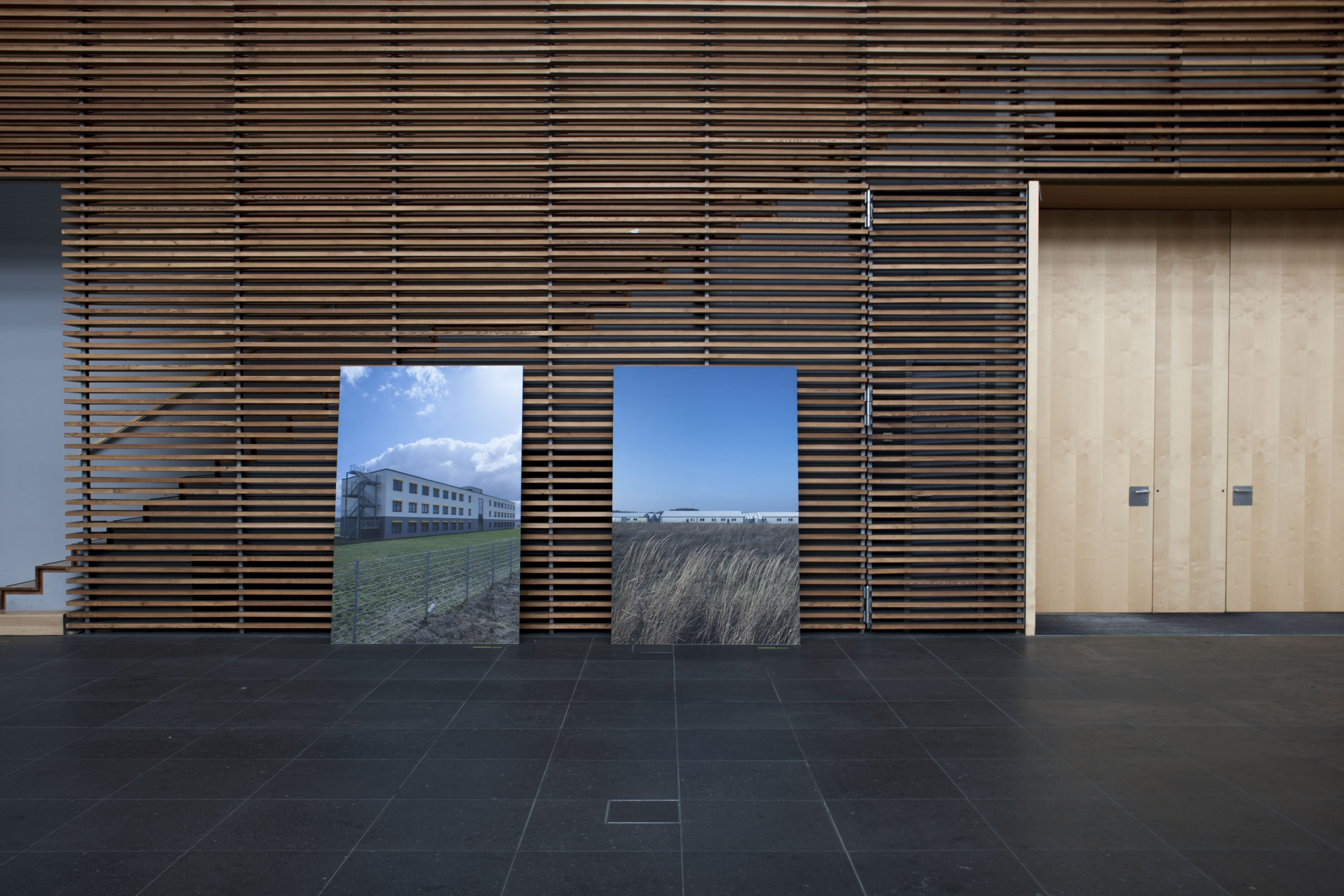 Two large photo panels lean against a slatted wooden wall in a foyer: one shows a white multi-storey building behind a fence, the other container buildings behind a field of grass. Denied Refuge / Verwehrte Zuflucht (2019) by Nickii Schamborski.