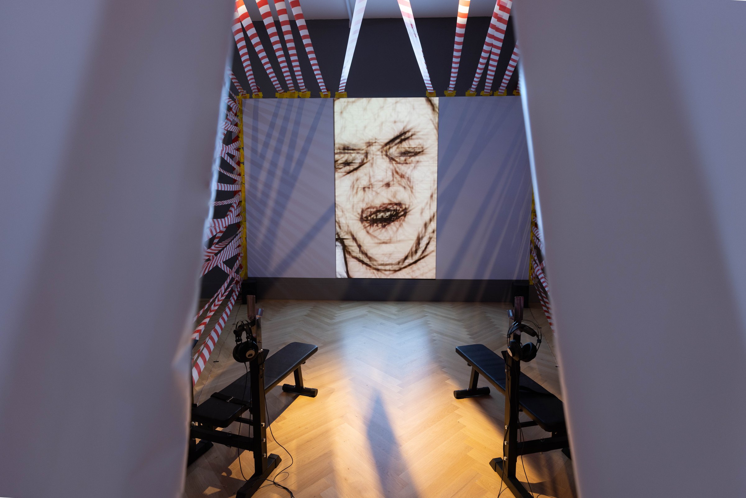 View from above into the installation: a room wrapped in red-and-white barrier tape with a projection of a grimacing face drawn in outlines, and weight benches with headphones in front. Among Us / Unter Uns (2021) by Nickii Schamborski.