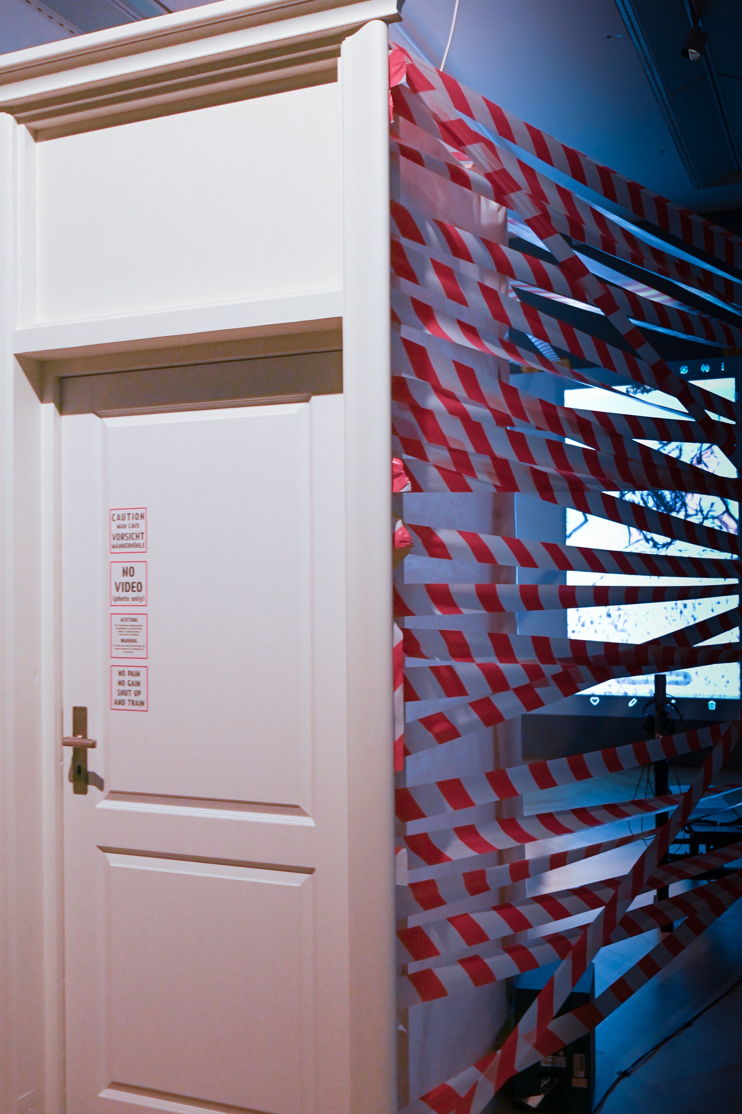 A white door in a free-standing frame; behind it, walls woven from red-and-white barrier tape with a projection visible through the gaps. Among Us / Unter Uns (2021) by Nickii Schamborski.