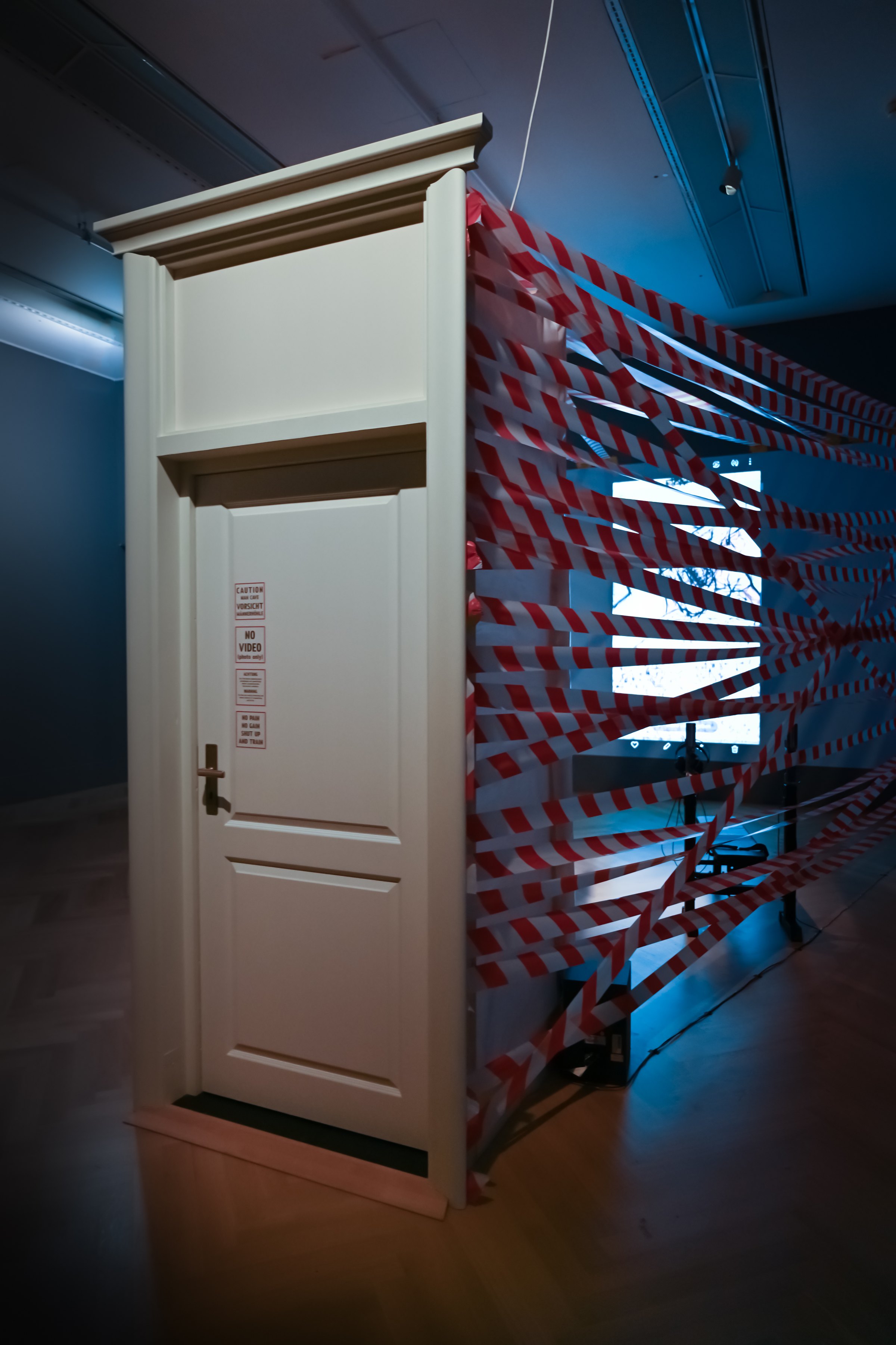 The installation in a dark exhibition hall: a free-standing white door with a room made of red-and-white barrier tape behind it. Among Us / Unter Uns (2021) by Nickii Schamborski.