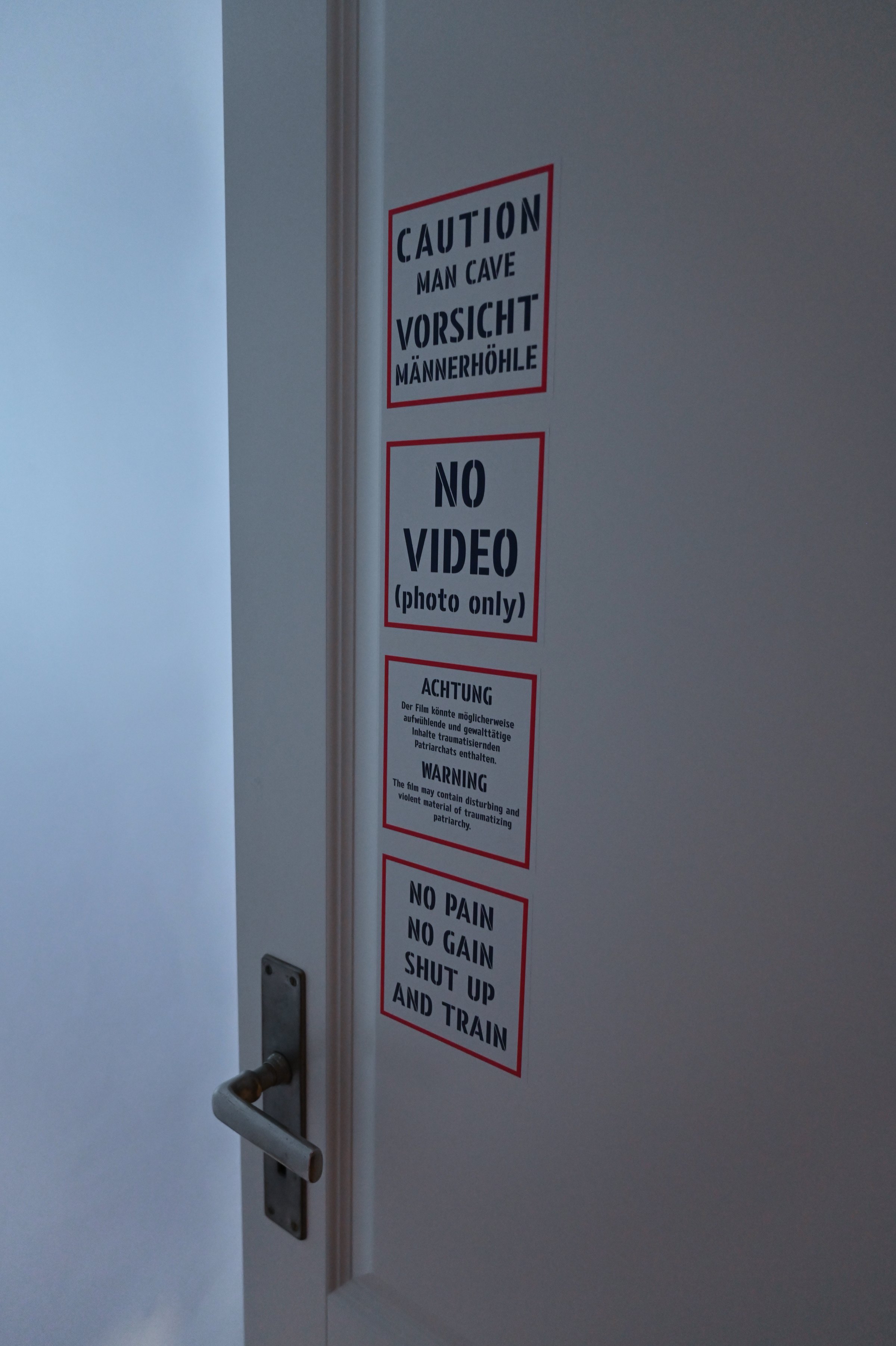 Close-up of the door with warning signs: 'Caution man cave / Vorsicht Männerhöhle', 'No video (photo only)', 'No pain no gain, shut up and train'. Among Us / Unter Uns (2021) by Nickii Schamborski.