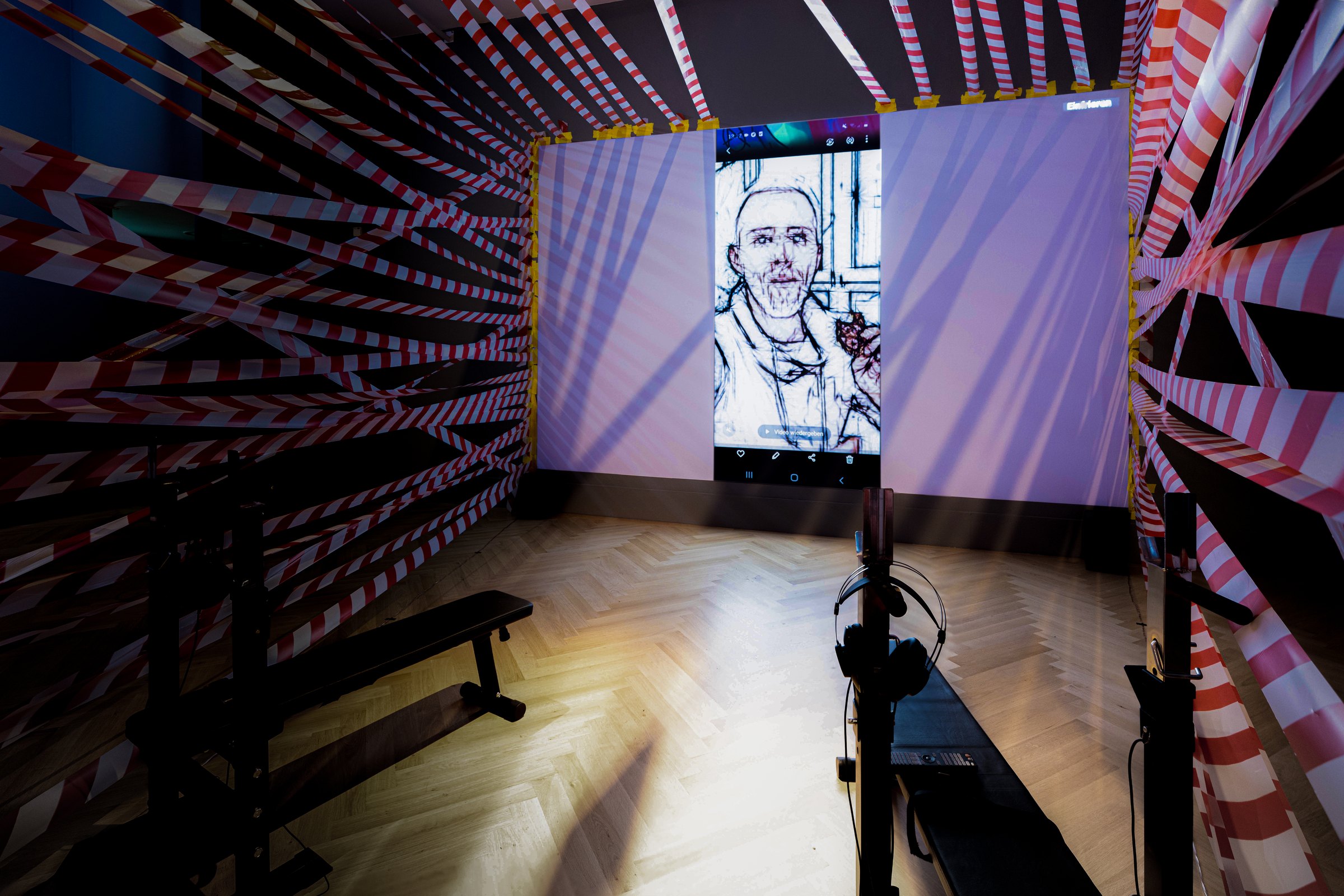 Inside the installation: a vertical phone-format projection showing a line drawing of a bearded man, with weight benches and headphones in the foreground. Among Us / Unter Uns (2021) by Nickii Schamborski.