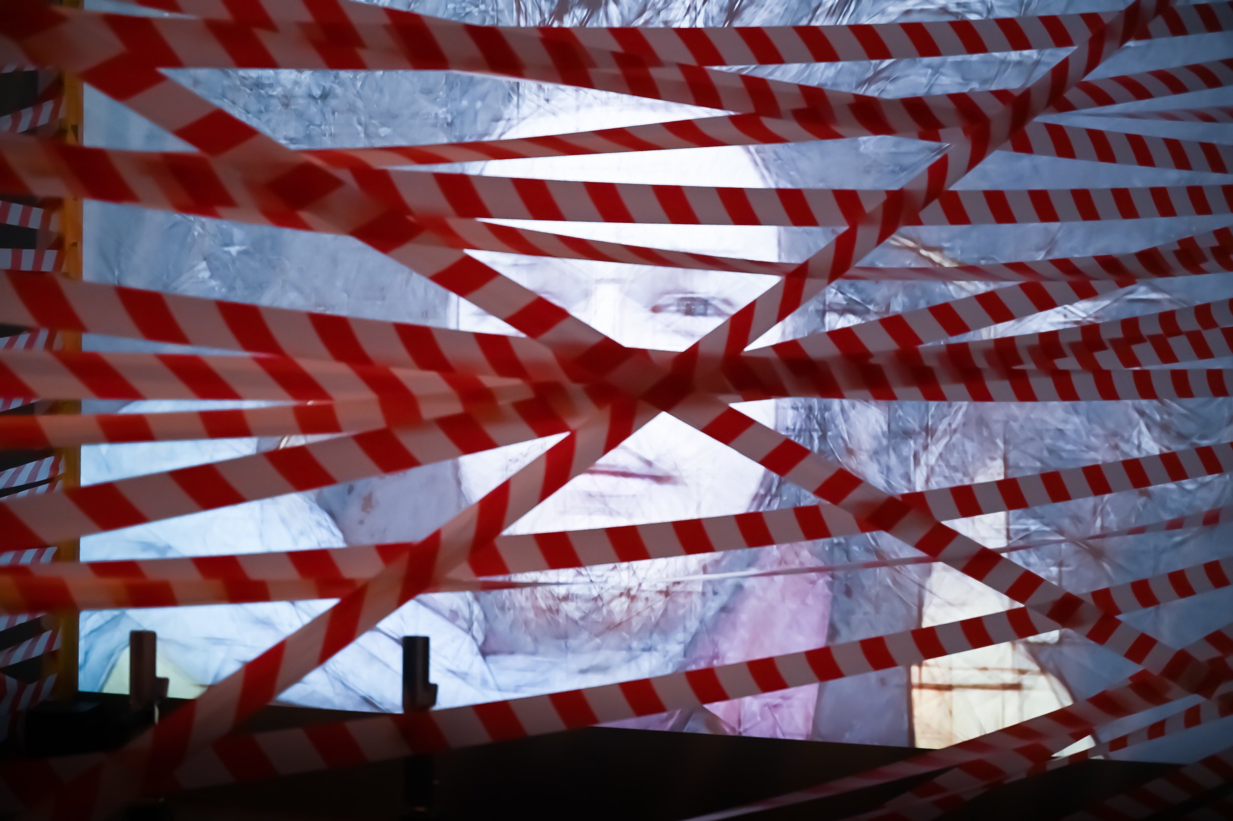 The projection seen from outside through crossing strips of red-and-white barrier tape. Among Us / Unter Uns (2021) by Nickii Schamborski.