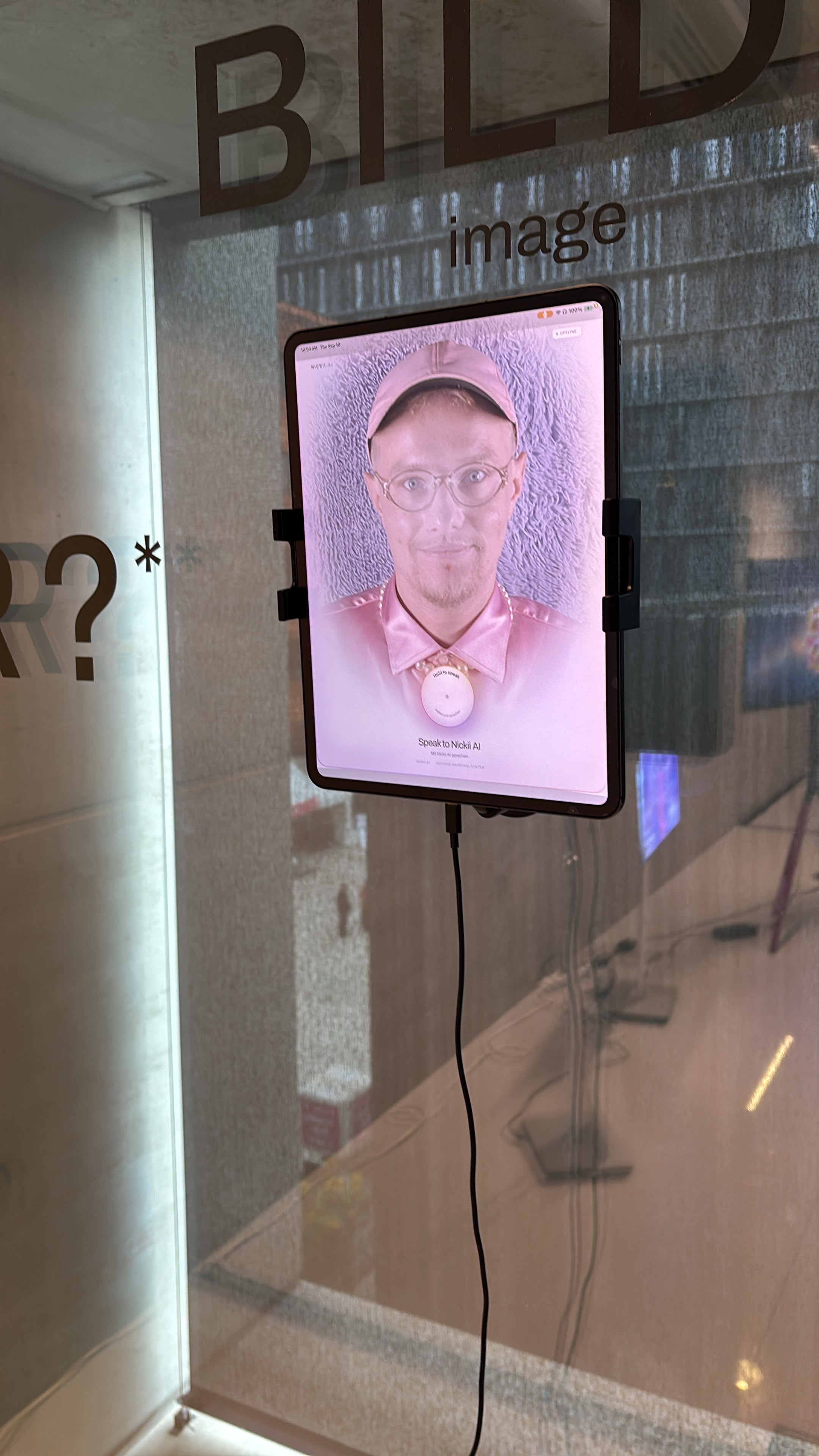A tablet mounted on a glass wall shows a face with a pink cap and glasses on a pink background, above the words "Speak to Nickii AI". Performance: Nickii AI (2026) by Nickii Schamborski.