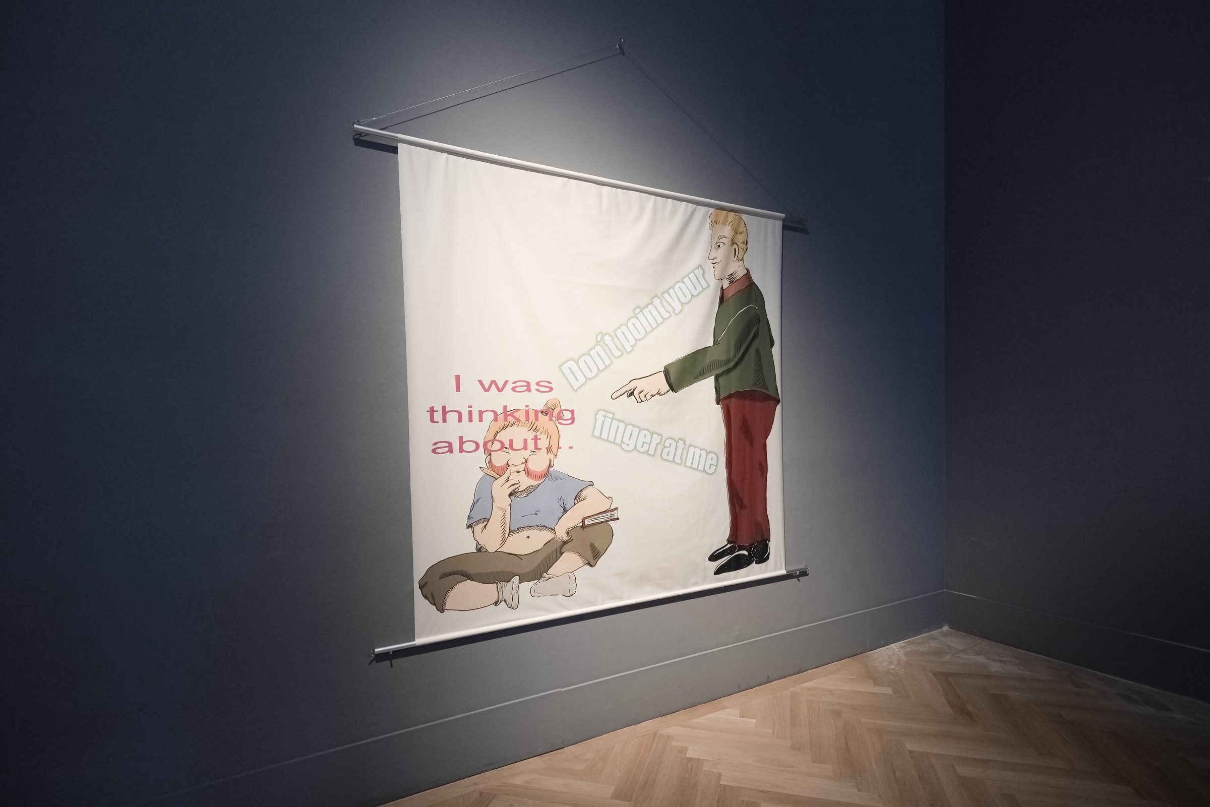 Fabric print hanging from an aluminium tube on a dark blue wall: a cartoon adult points at a crying child sitting on the ground; the texts read 'I was thinking about…' and 'Don't point your finger at me'. MeMärchen (2021) by Nickii Schamborski.