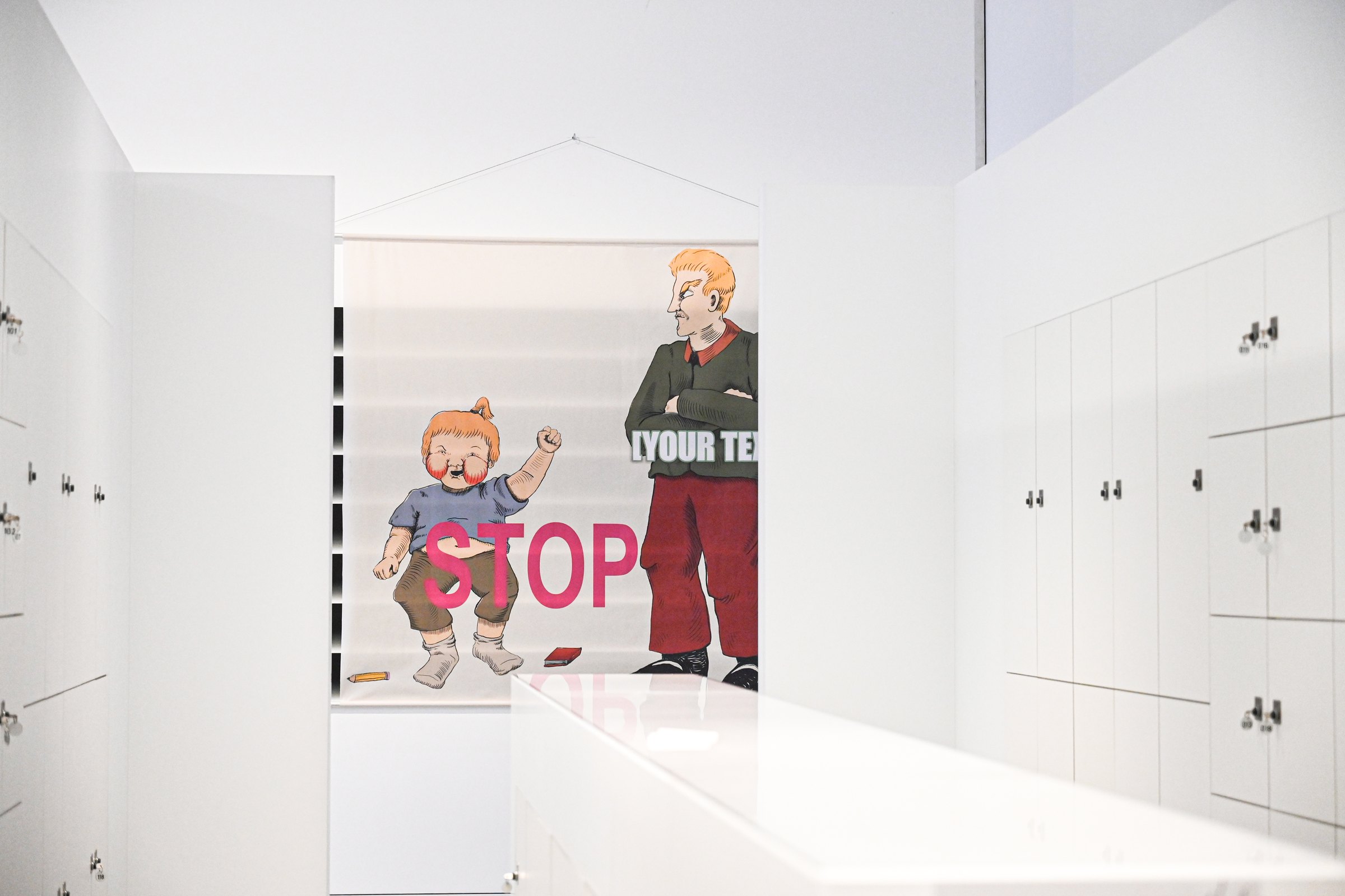 Fabric print at the end of a white locker corridor: a cartoon child raises a fist next to an adult with crossed arms; large pink text reads 'STOP'. MeMärchen (2021) by Nickii Schamborski.