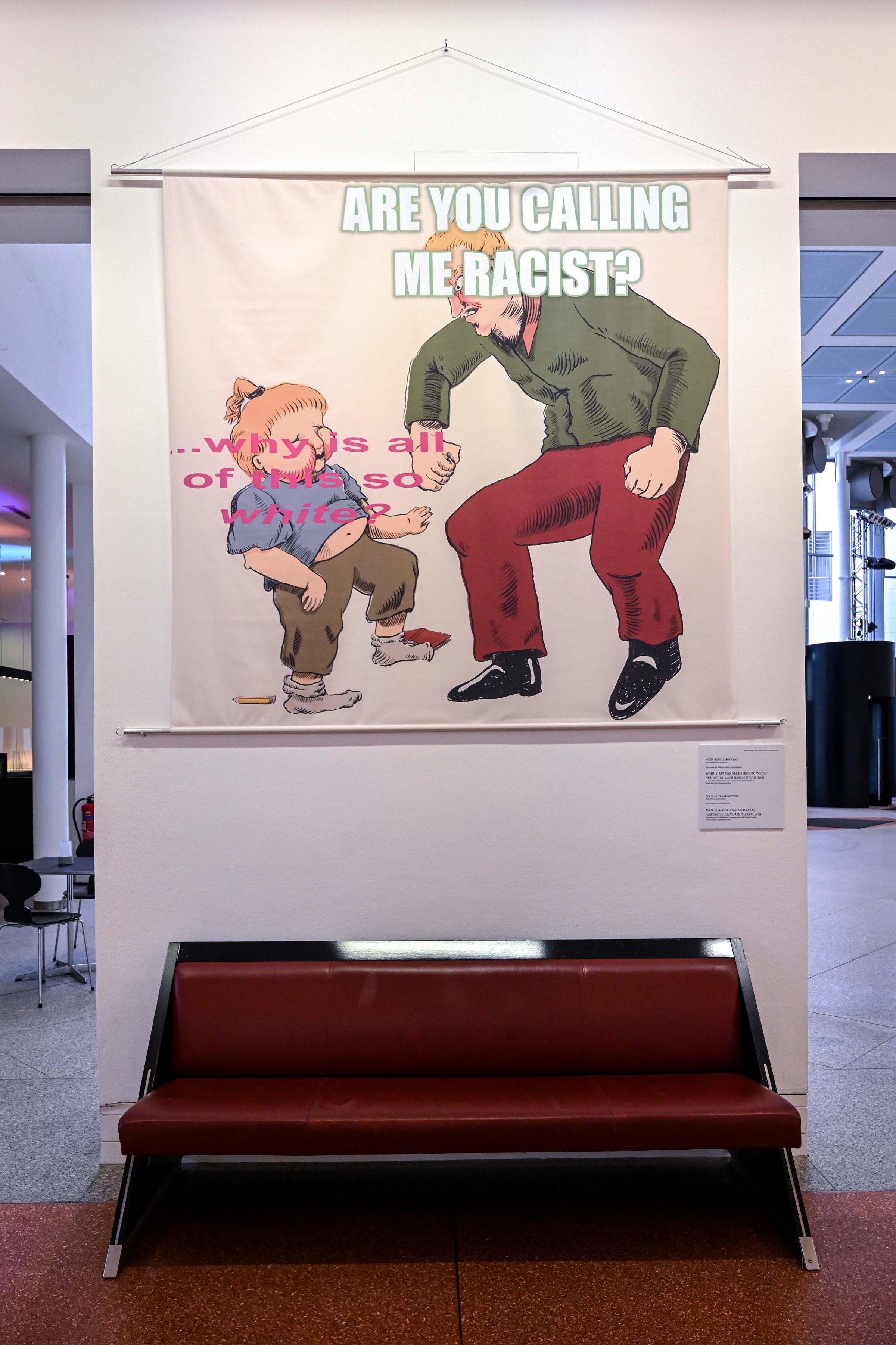 Fabric print above a red bench: a cartoon adult bends over a crying child; the text reads 'Are you calling me racist?'. MeMärchen (2021) by Nickii Schamborski.