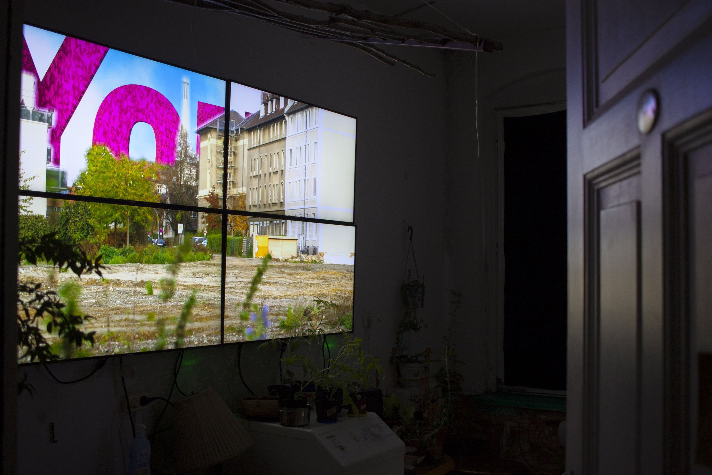 Installation view: four wall-mounted monitors together show an empty urban plot with large pink letters, in a dark room with plants. Create Your Surrounding! (2017) by Nickii Schamborski.