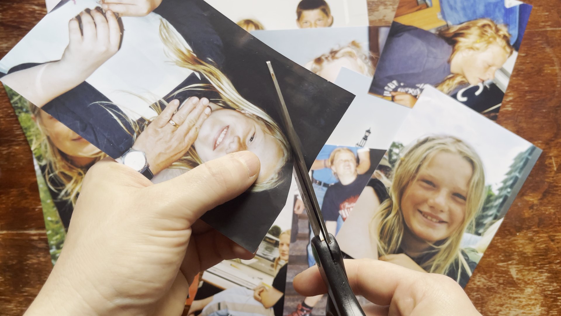 Hands cut a printed childhood photograph with scissors above a pile of other family photos. CyberCindy (2025) by Nickii Schamborski.