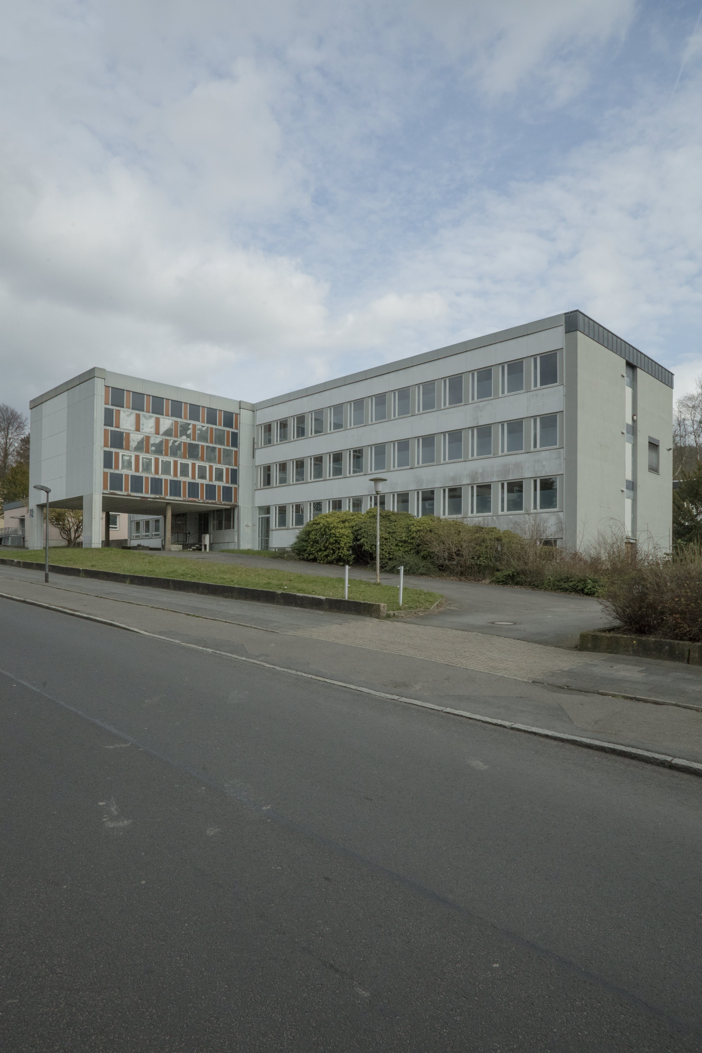 Photograph: a grey three-storey office building with coloured panels beside an empty road. Denied Refuge / Verwehrte Zuflucht (2019) by Nickii Schamborski.