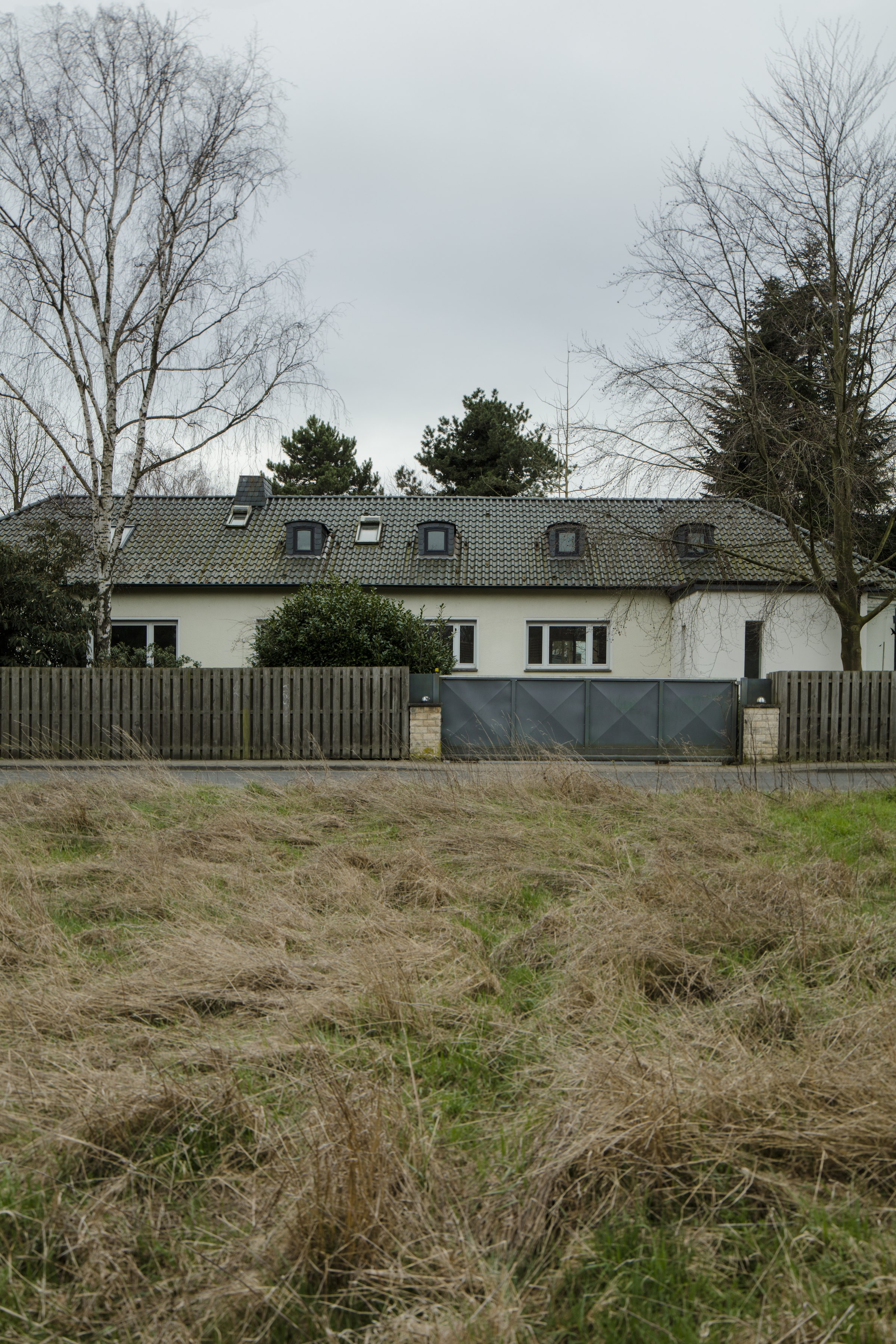 Photograph: a low white house behind a wooden fence, with an overgrown field of dry grass in the foreground. Denied Refuge / Verwehrte Zuflucht (2019) by Nickii Schamborski.
