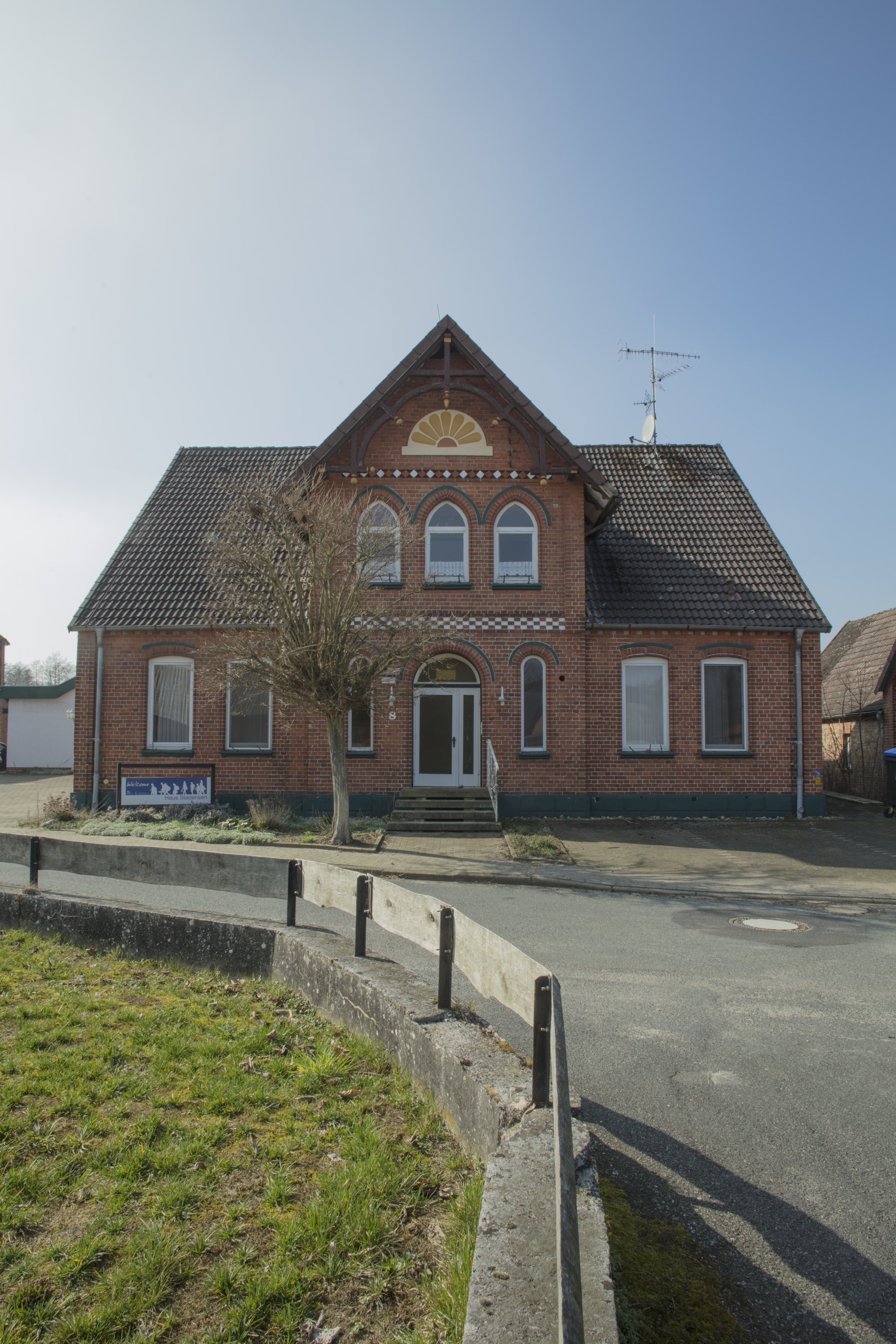 Photograph: a red-brick house with arched windows and a gabled roof at a street corner. Denied Refuge / Verwehrte Zuflucht (2019) by Nickii Schamborski.