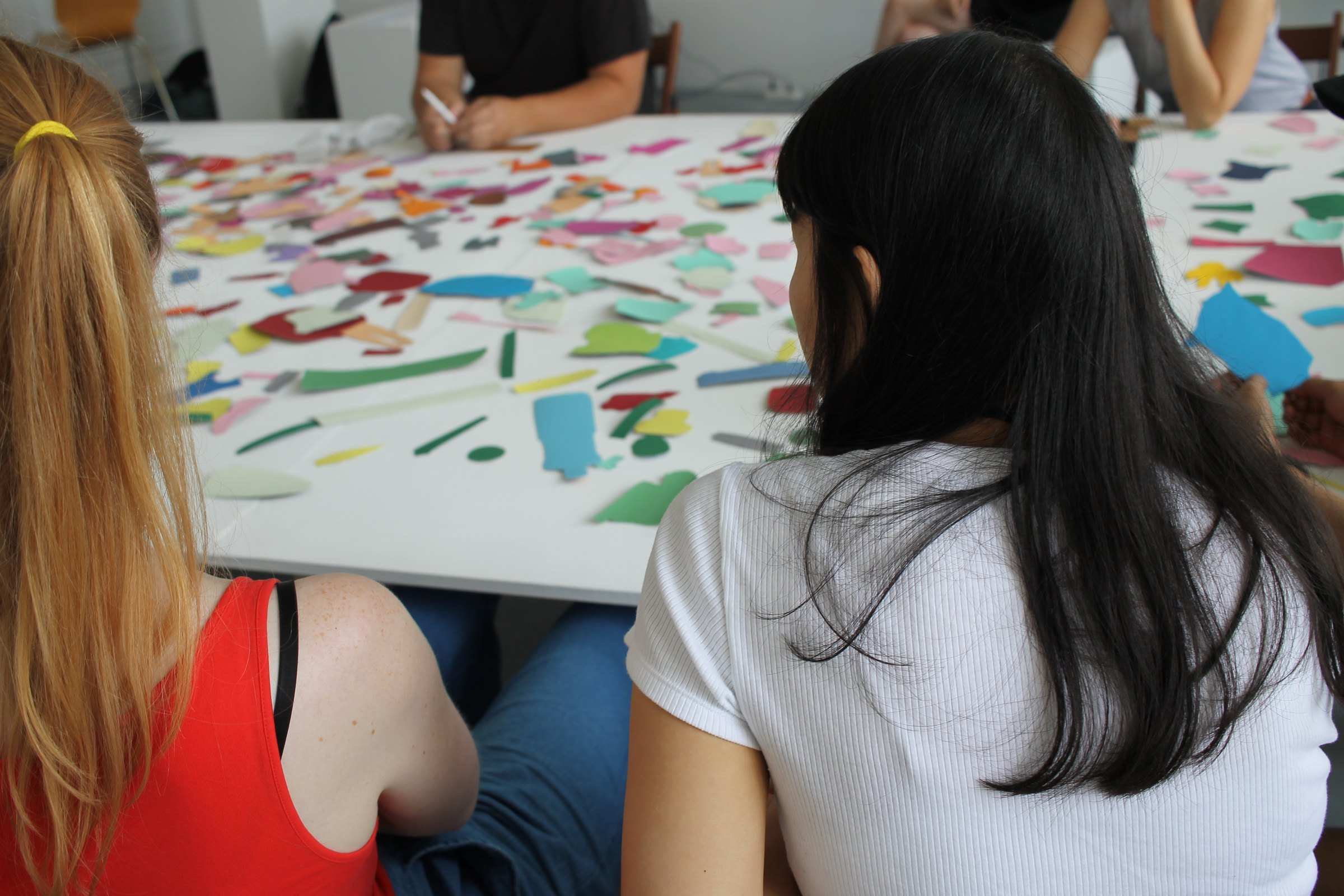 Participants seen from behind sit around a table covered with small colourful paper shapes. Entangled Responsibility (2016) by Nickii Schamborski.