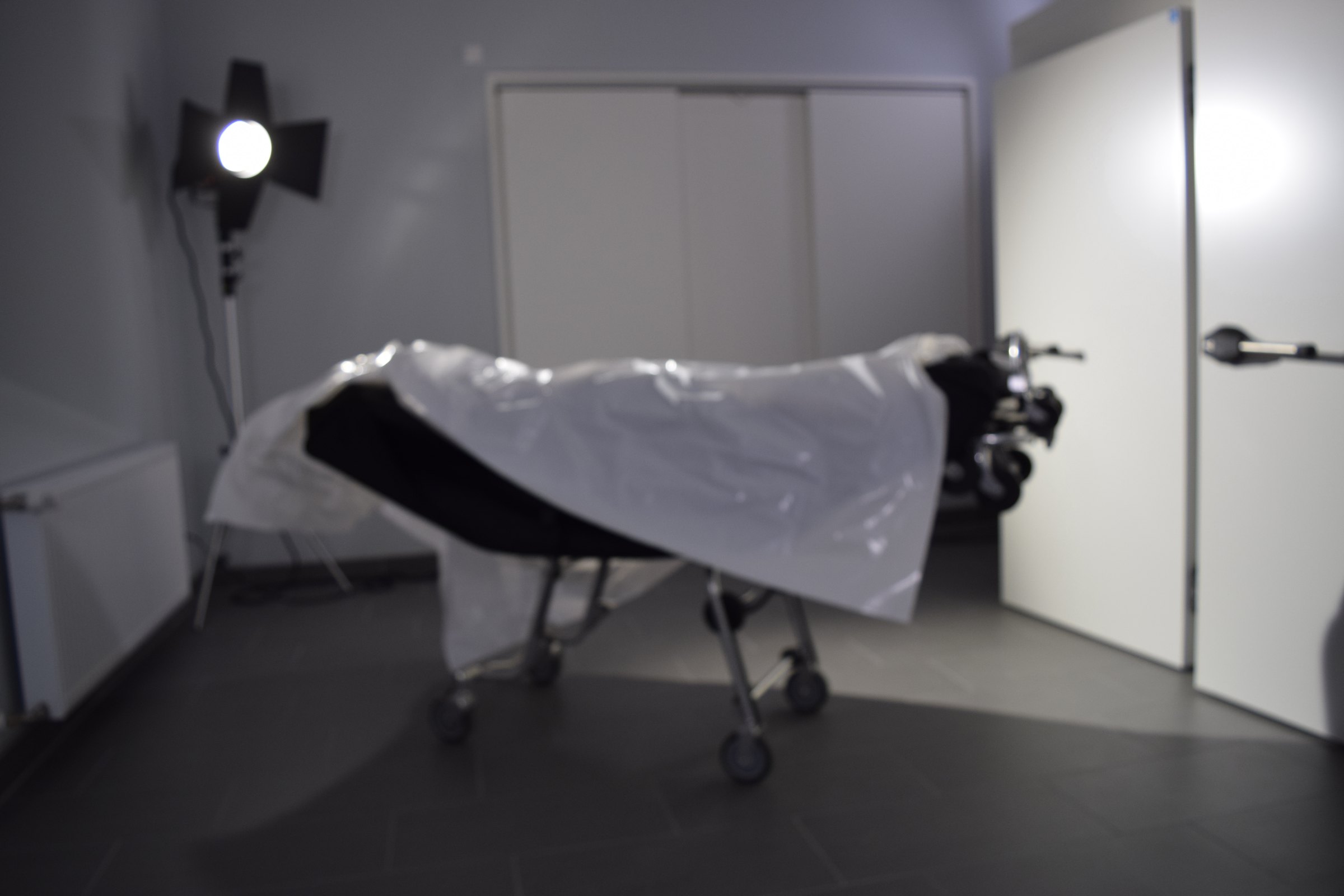 Set photo: a stretcher covered with a plastic sheet stands in a grey room, lit by a film light. Familycare (2017/2019) by Nickii Schamborski.