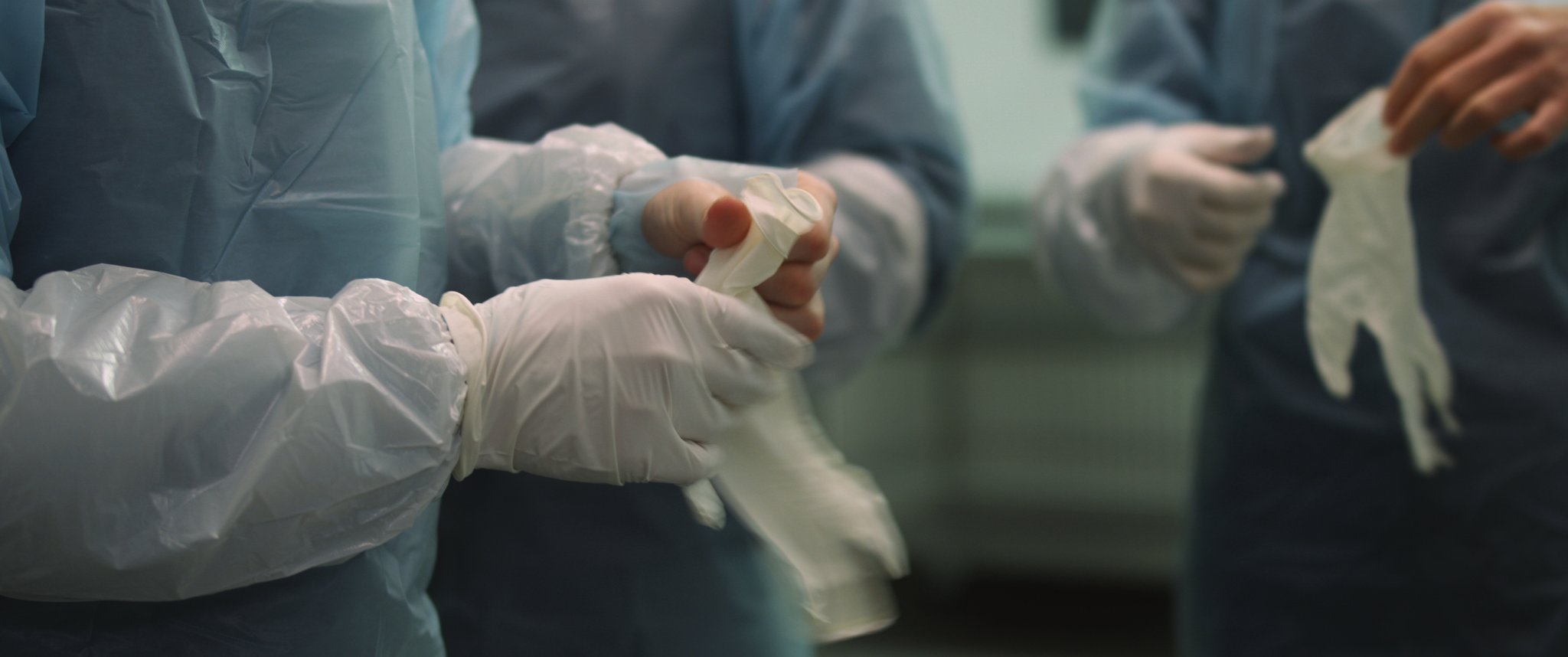 Video still: close-up of hands in disposable gowns pulling on white latex gloves. Familycare (2017/2019) by Nickii Schamborski.
