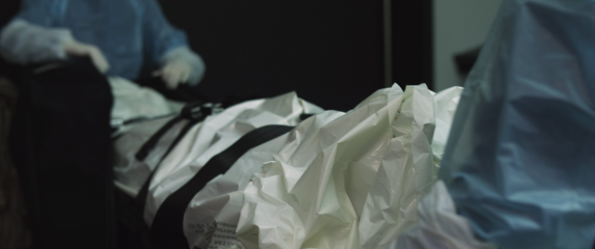 Video still: gloved hands open a white body bag with black straps. Familycare (2017/2019) by Nickii Schamborski.