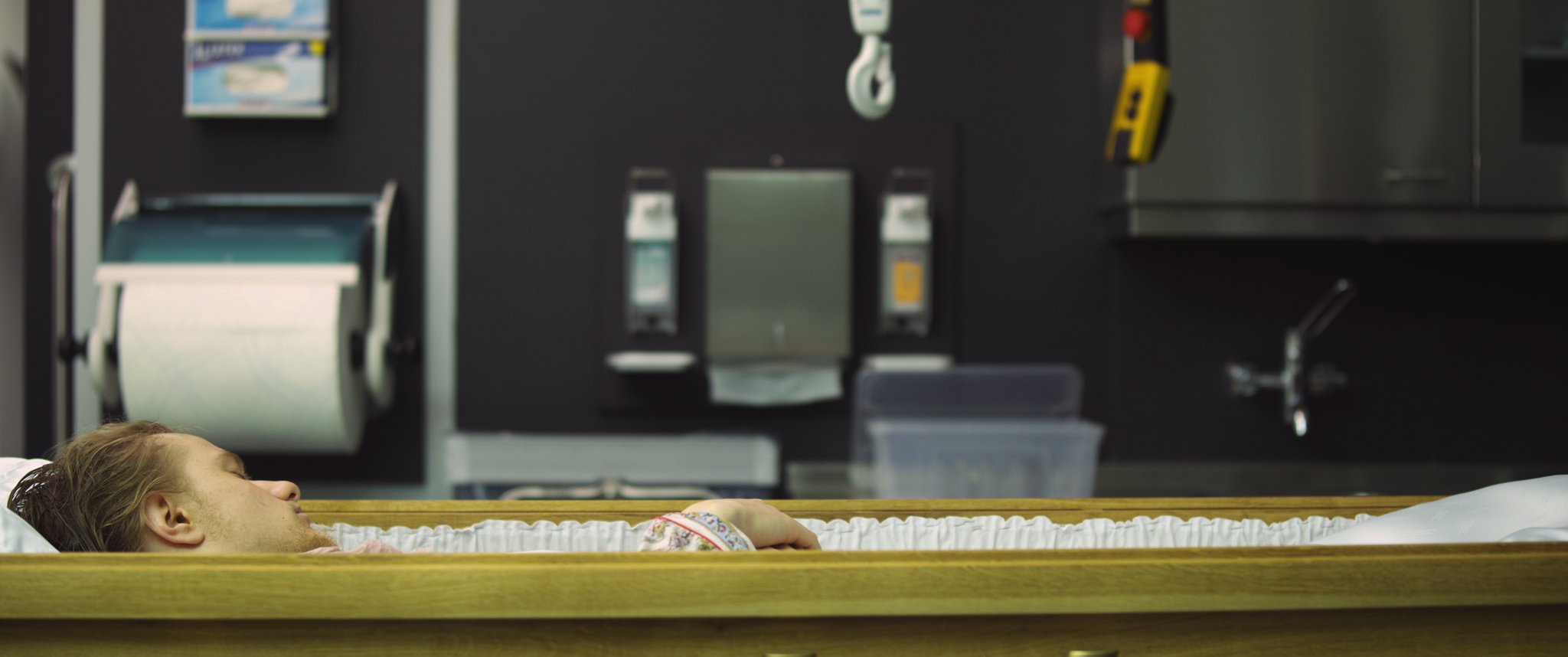 Video still: a person lies in an open wooden coffin, seen from the side, in a dark tiled preparation room. Familycare (2017/2019) by Nickii Schamborski.