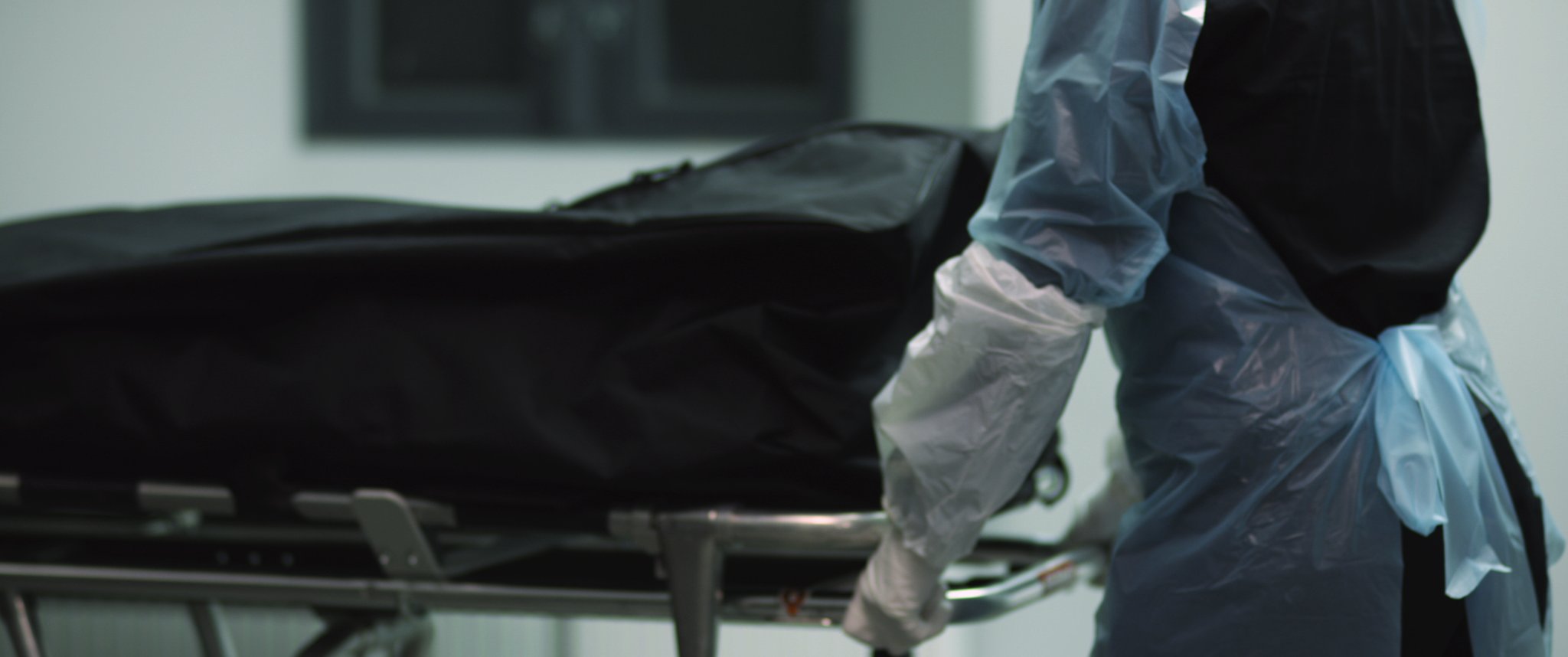 Video still: a person in a blue disposable gown and gloves pushes a stretcher carrying a black body bag. Familycare (2017/2019) by Nickii Schamborski.