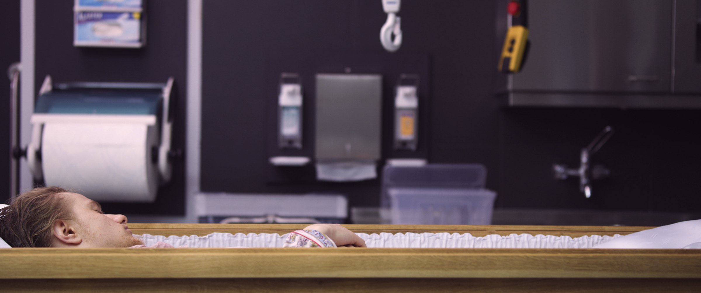 Video still in cinemascope: a person lies with closed eyes in a wooden coffin, seen in profile, in a tiled preparation room with a paper-towel dispenser and a sink. Familycare (2017/2019) by Nickii Schamborski.