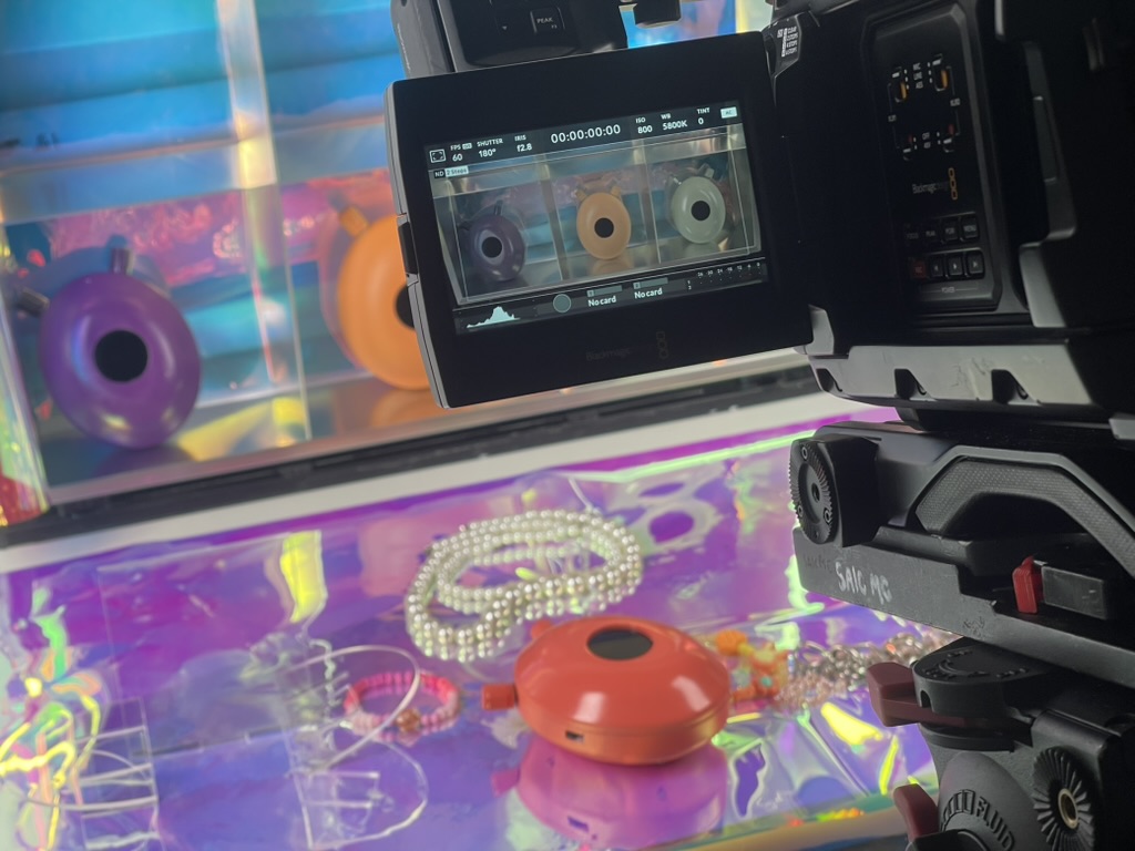 A camera films an orange device lying on iridescent foil with pearl necklaces; the camera monitor shows three devices in a row. Funke (2026) by Nickii Schamborski.