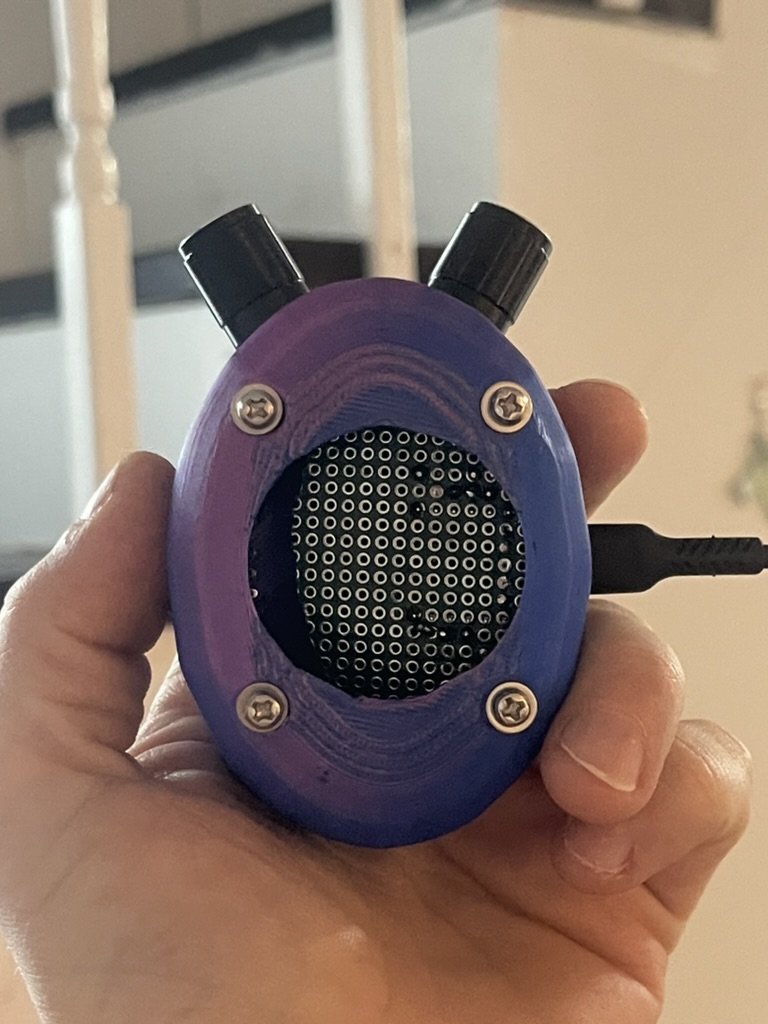 A hand holds an egg-shaped purple 3D-printed device with two black rotary knobs and a round opening showing a perforated circuit board. G-Spirit (2026) by Nickii Schamborski.