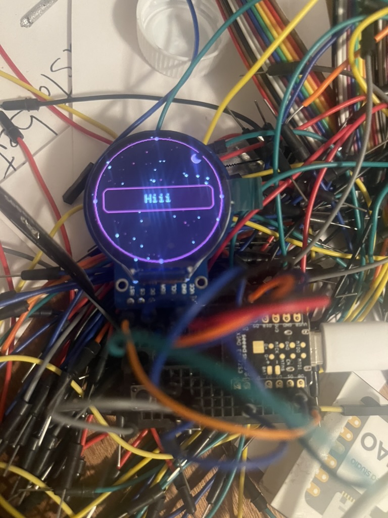 Prototype: a round display glowing purple with a blue bar, amid a tangle of jumper wires on a breadboard. G-Spirit (2026) by Nickii Schamborski.