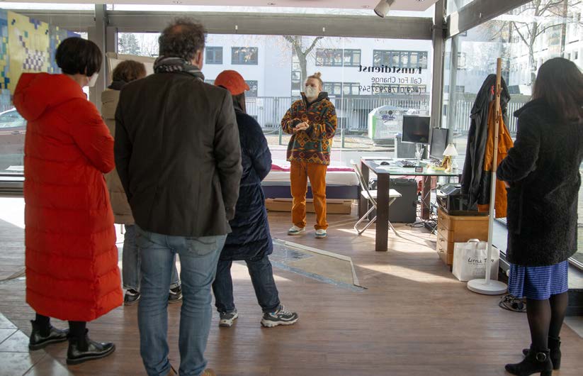 Visitors in winter coats stand in a glass-fronted shop space and listen to a person in a patterned fleece jacket and face mask; mirrored lettering 'Kunstdienst' on the window. Kunstdienst (2022) by Nickii Schamborski.