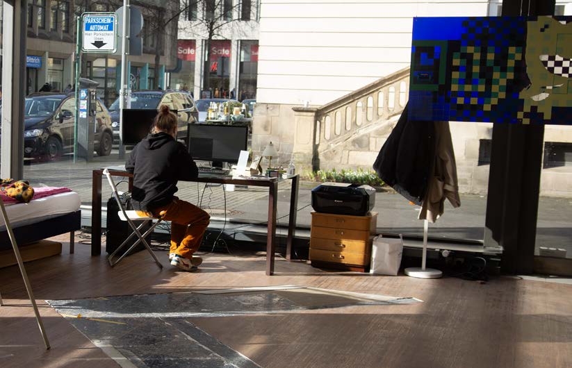 A person sits at a desk with a monitor inside a glass-fronted shop space; a pixelated blue and yellow banner hangs in the window, the street visible outside. Kunstdienst (2022) by Nickii Schamborski.