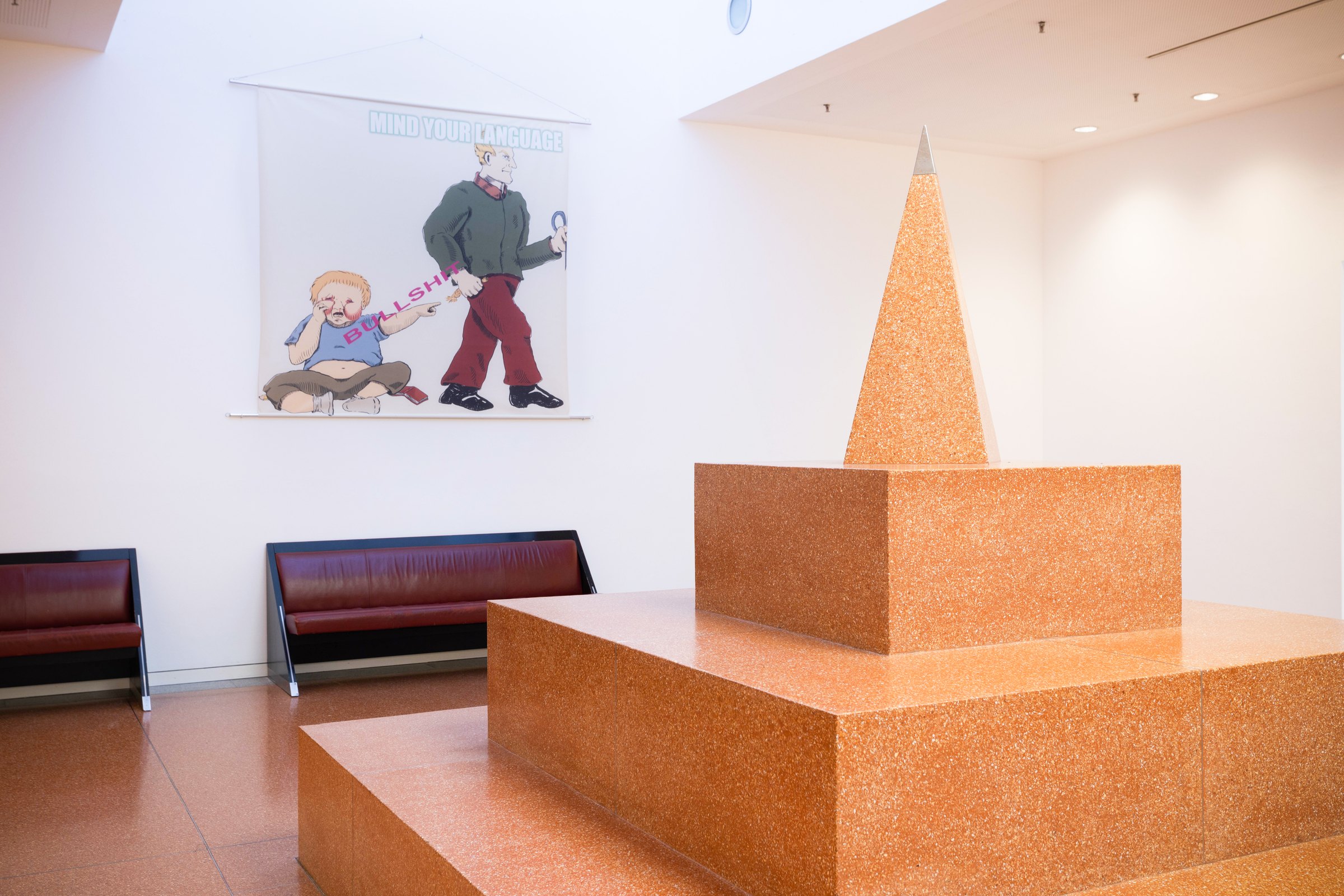 Fabric print on the wall of a foyer with benches and an orange stone pyramid: a cartoon adult with a cane stands over a sitting child; the text reads 'Mind your language!'. MeMärchen (2021) by Nickii Schamborski.