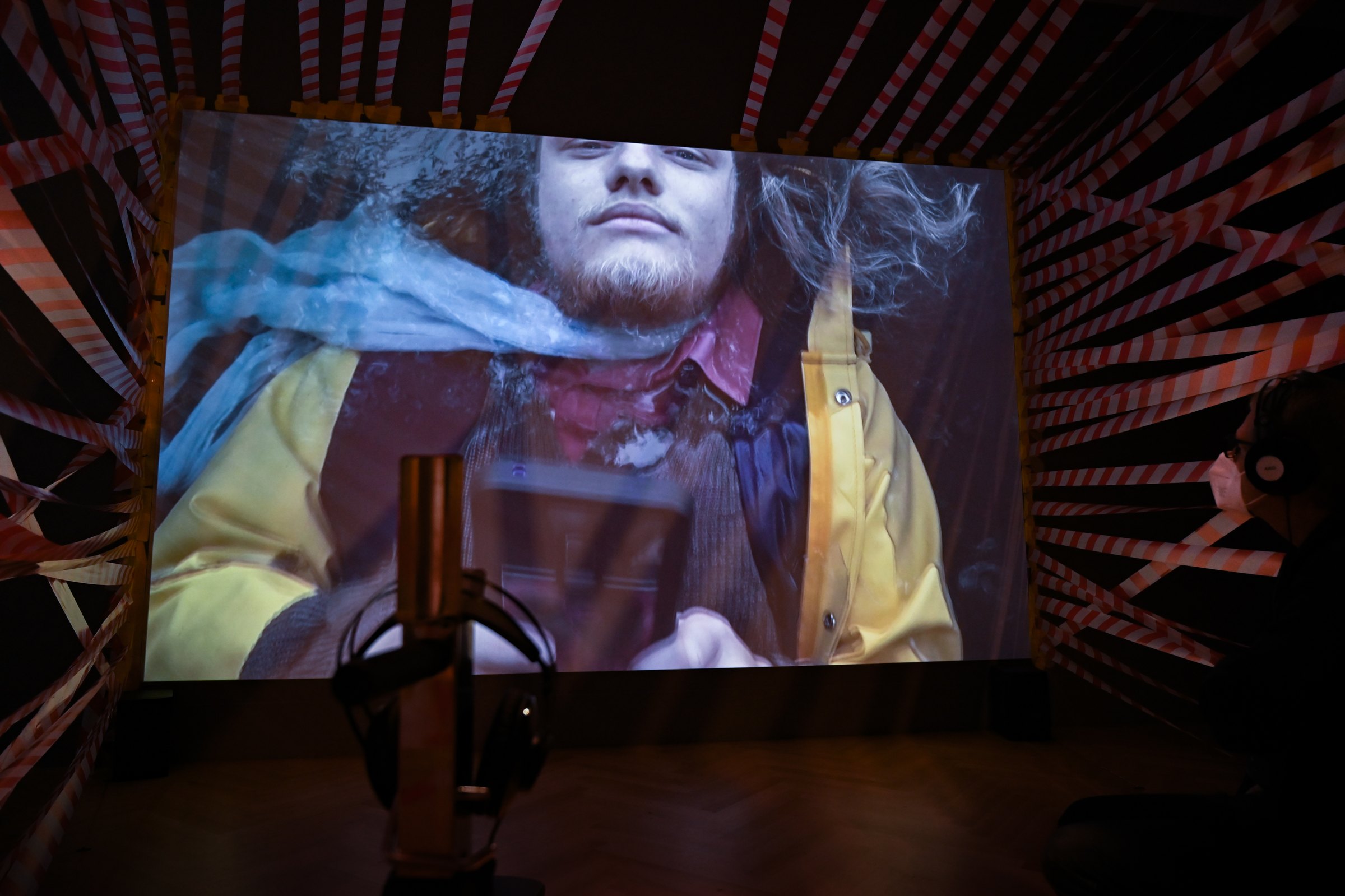Inside the installation: a large projection of a bearded person with long hair seen from below, surrounded by walls of barrier tape; a visitor with headphones sits on the right. Among Us / Unter Uns (2021) by Nickii Schamborski.