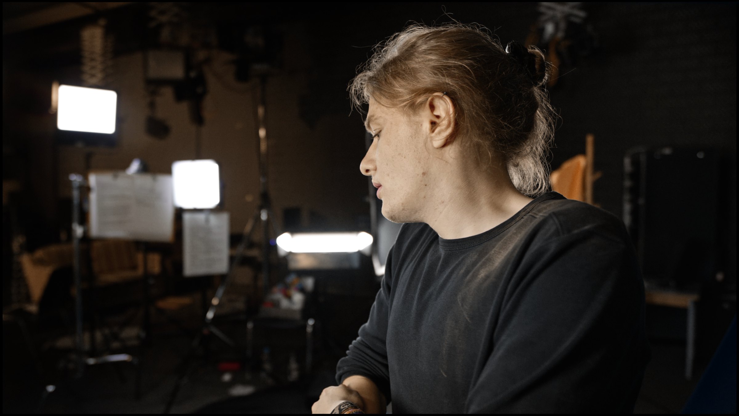 A person with hair tied back, in a dark sweatshirt, looks down to the side in a dark film studio with light panels in the background. Systemchange (2020) by Nickii Schamborski.