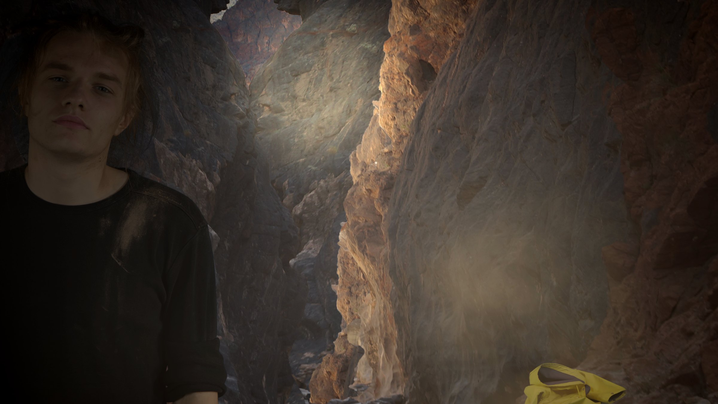 A person in a dark T-shirt stands in a dim rocky cave lit by a shaft of light; a yellow raincoat lies on the ground. Systemchange (2020) by Nickii Schamborski.