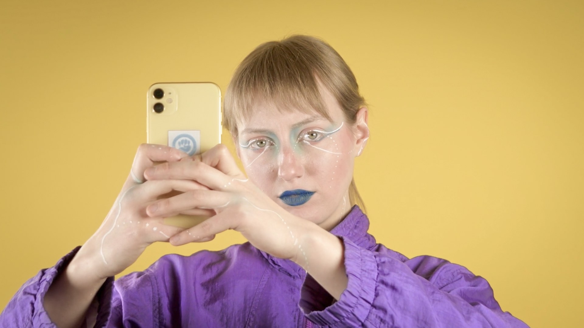 A person with blue lipstick and white graphic eye make-up, wearing a purple blouse, holds a yellow smartphone up to their face against a yellow background. Anastasiia (2024) by Nickii Schamborski.