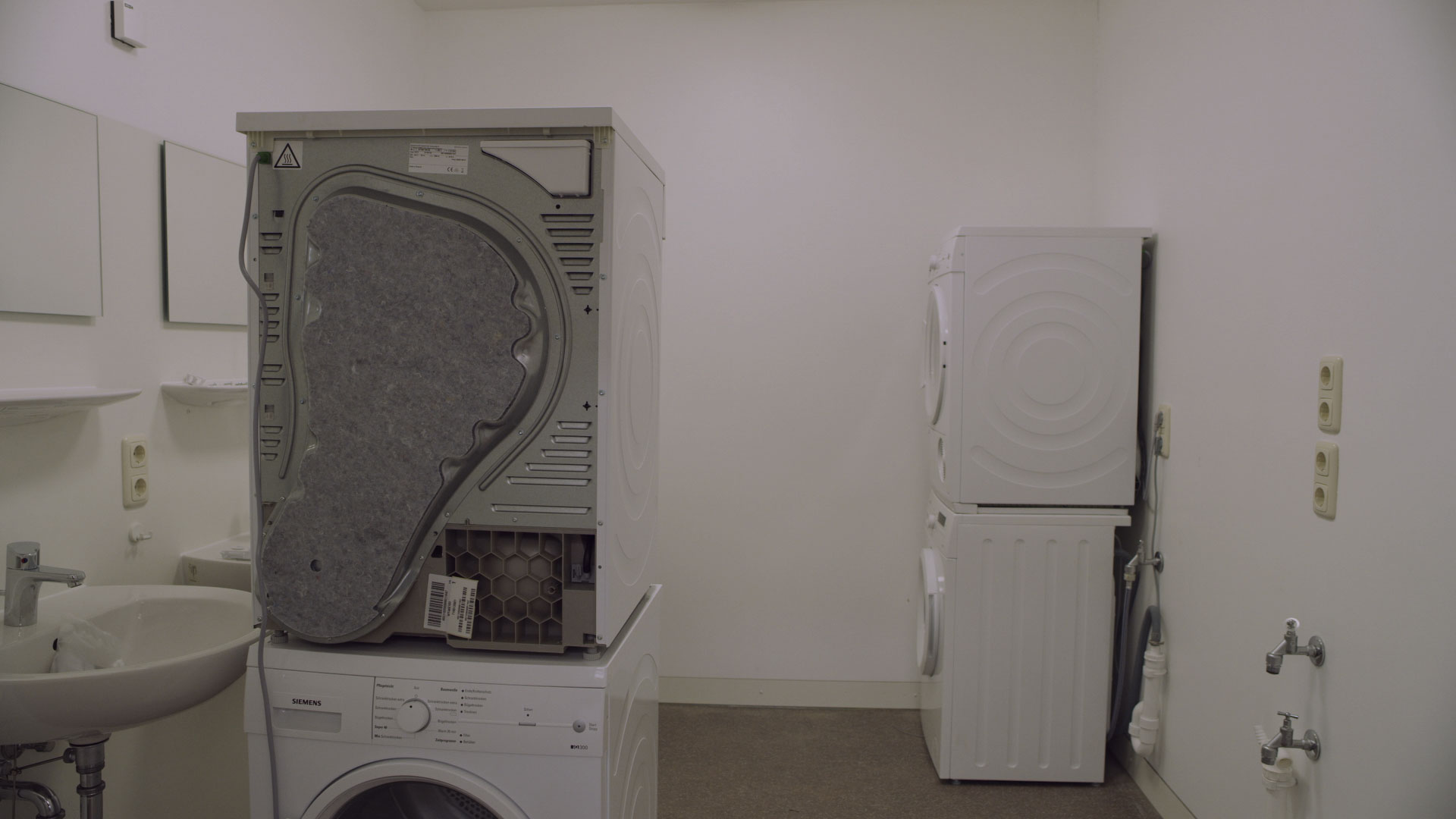 Video still: a bare white room with washing machines and sinks, one machine turned to show its back. Unused Capacity (2018) by Nickii Schamborski.