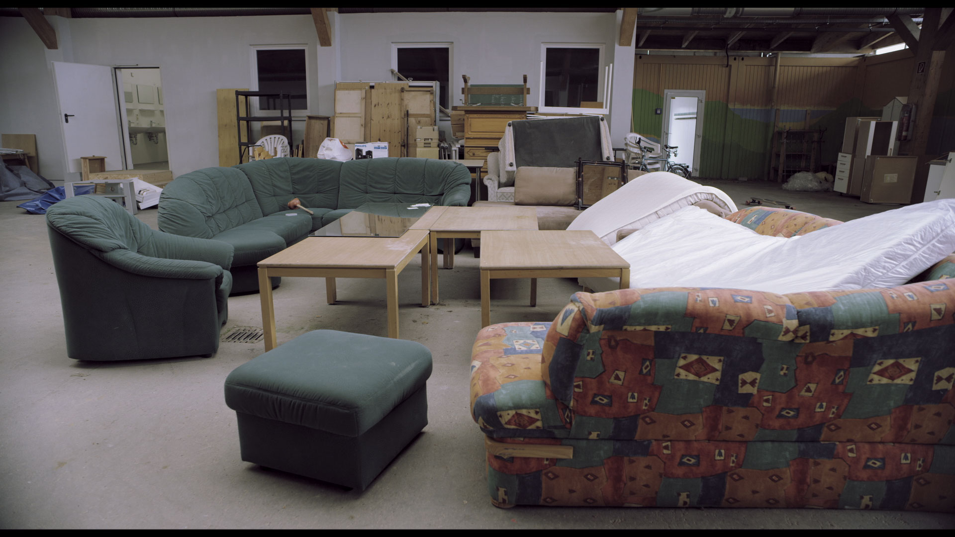 Video still: a storage hall full of unused sofas, armchairs, tables and mattresses. Unused Capacity (2018) by Nickii Schamborski.