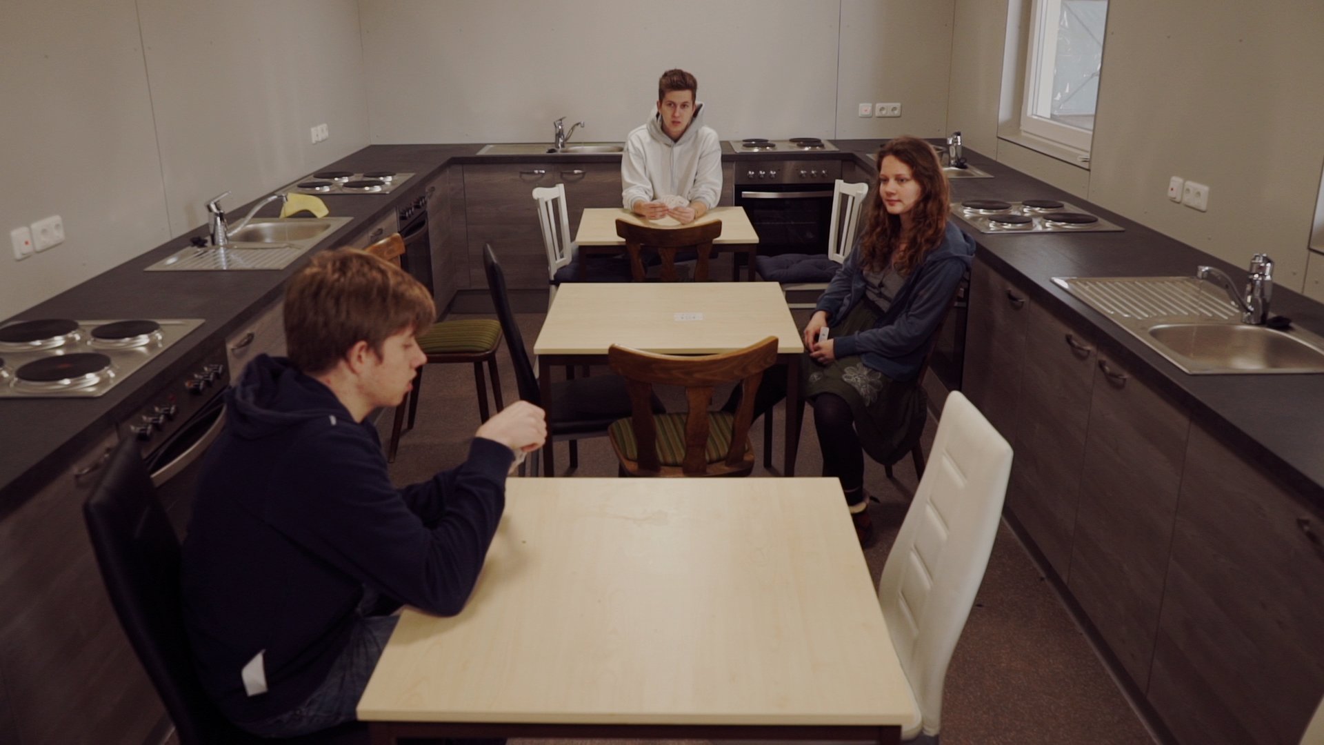 Video still: three young people sit at separate tables in a kitchen lined with hobs and sinks. Unused Capacity (2018) by Nickii Schamborski.